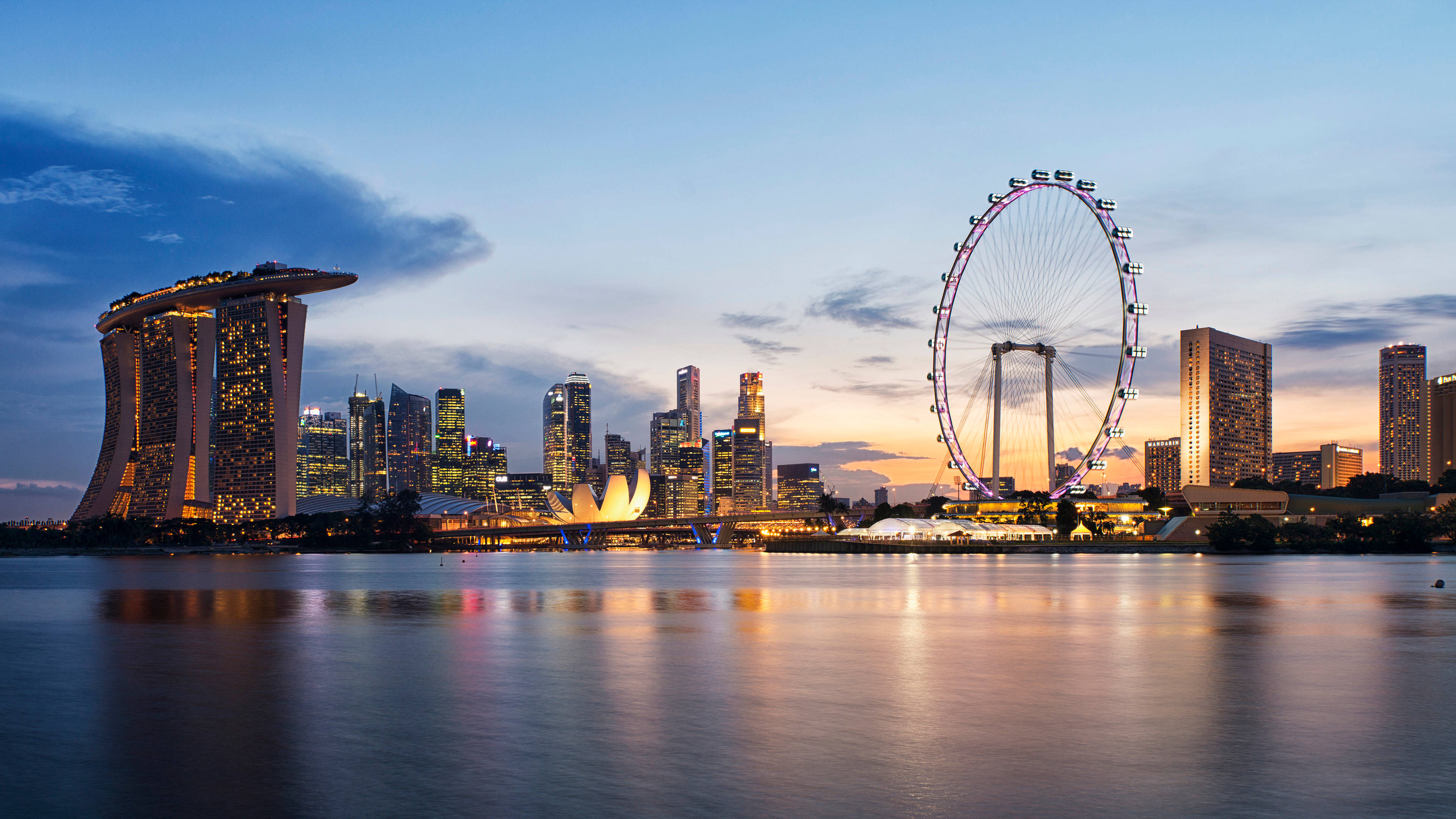 Singapore 桌布 壁紙 — 黃昏時分的新加坡天際線，濱海灣金沙和新加坡摩天輪在柔和藍橙色天空的映襯下閃耀，倒映在平靜的水面上。