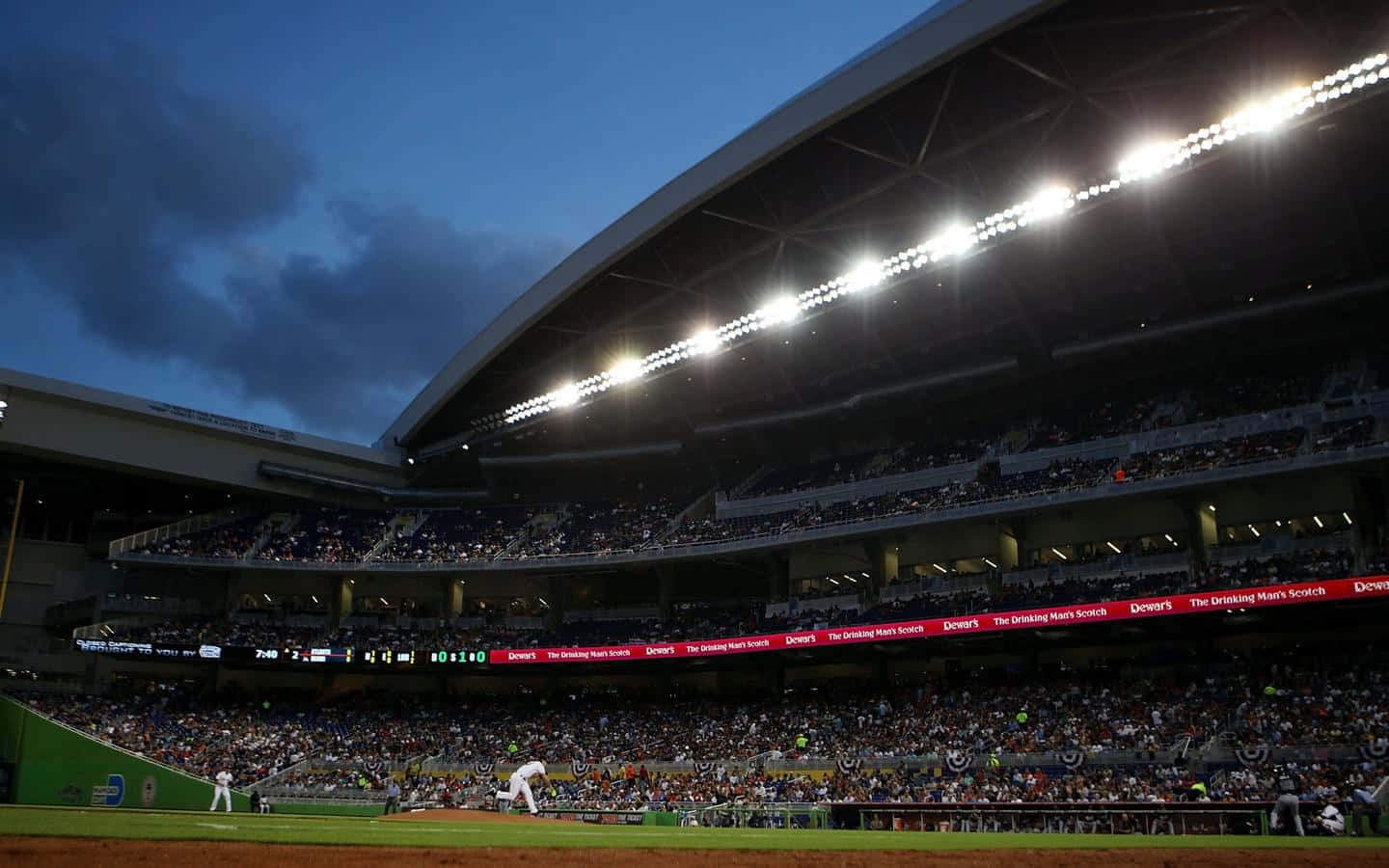 Baseball Stadium 桌布 壁紙 — 黃昏時分的棒球場，明亮的燈光照亮球場和擠滿觀眾的看台，天空多雲。