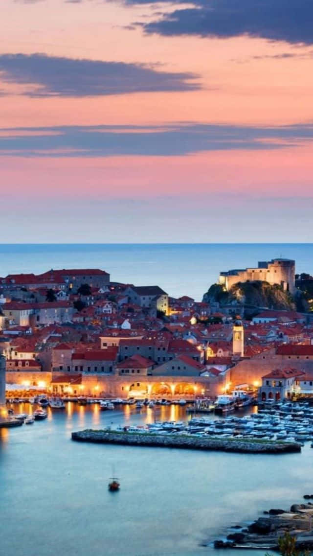 Croatia 桌布 壁紙 — 黃昏時分的沿海小鎮，溫暖的燈光映照在平靜的水面上，天空呈現粉紅色和藍色。