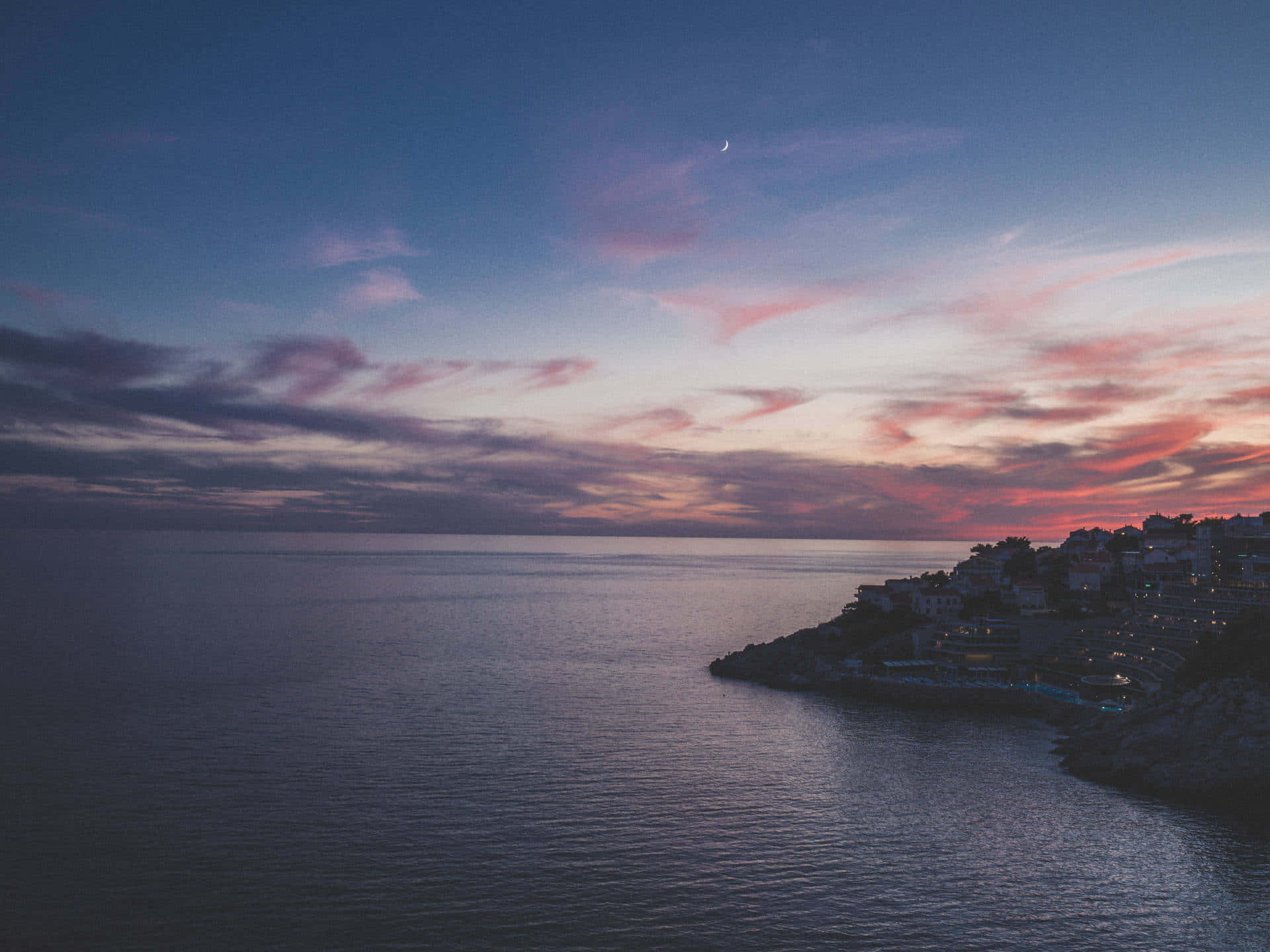 Clouds Aesthetics 桌布 壁紙 — 黃昏時分的沿海小鎮，粉紅色和紫色的雲層上方有一彎新月，映照在平靜的海面上。