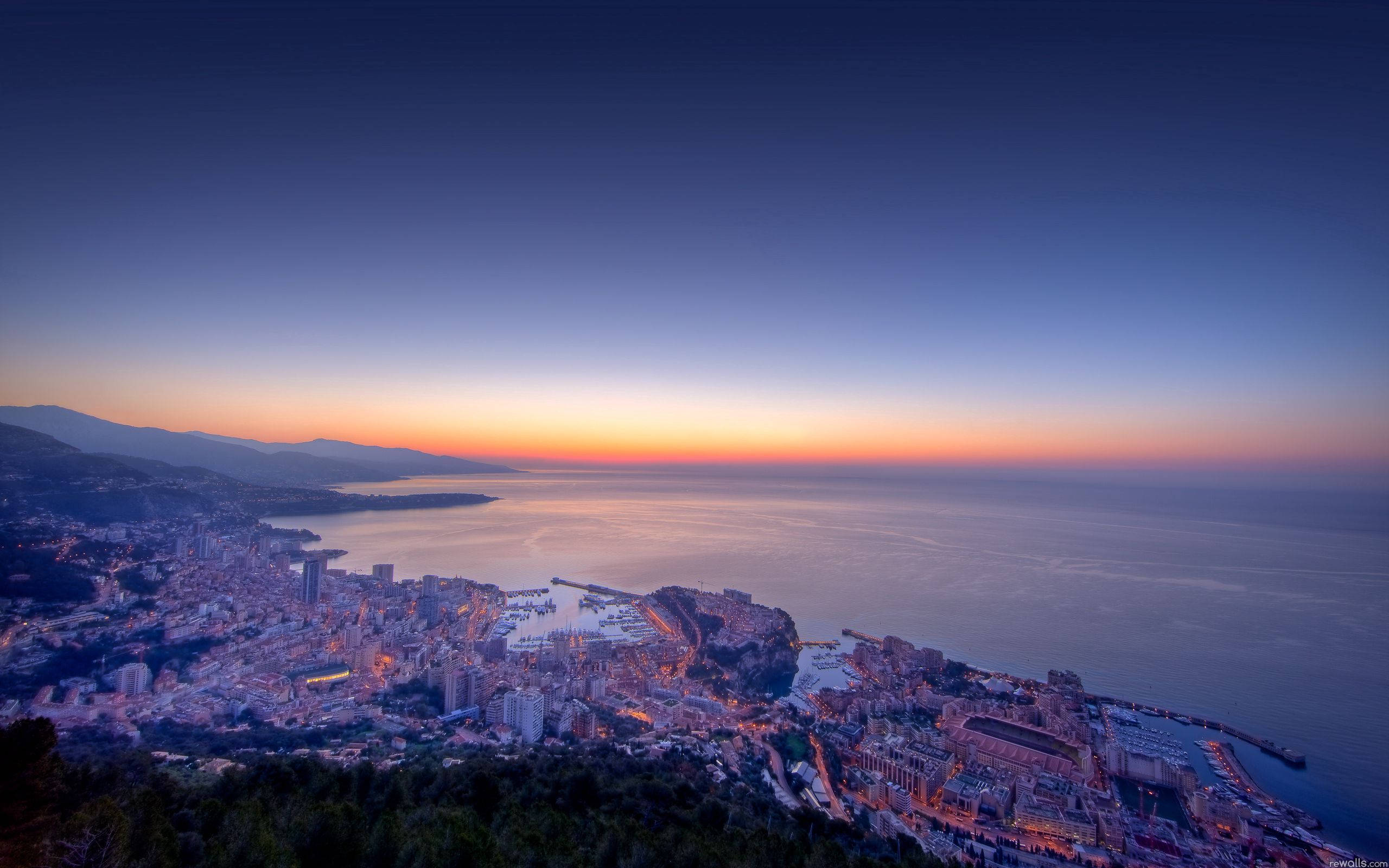 Monaco 桌布 壁紙 — 黃昏時分的海岸城市景觀，溫暖的橙色地平線映照在平靜的海面上，發光的建築依偎在山巒之間。