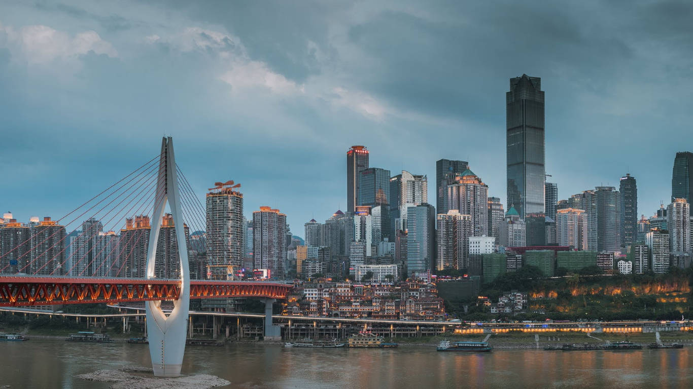 Chongqing 桌布 壁紙 — 黃昏時分的現代城市景觀，一座發光的紅色橋樑橫跨河流，摩天大樓倒映在水中。
