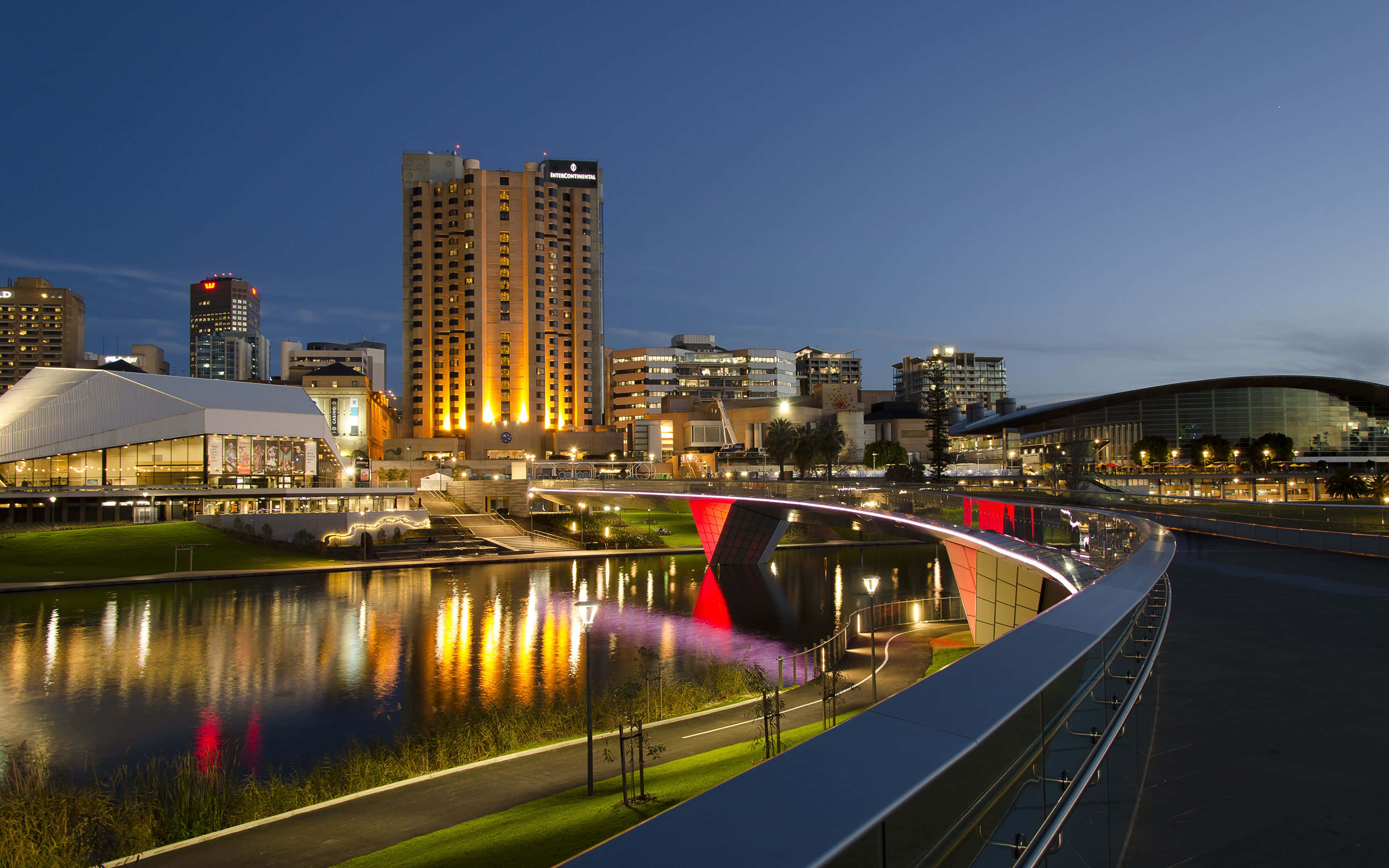 Adelaide 桌布 壁紙 — 黃昏時分的現代城市景觀，燈火通明的摩天大樓和一座在平靜河流上反射溫暖光芒的發光紅色橋樑。