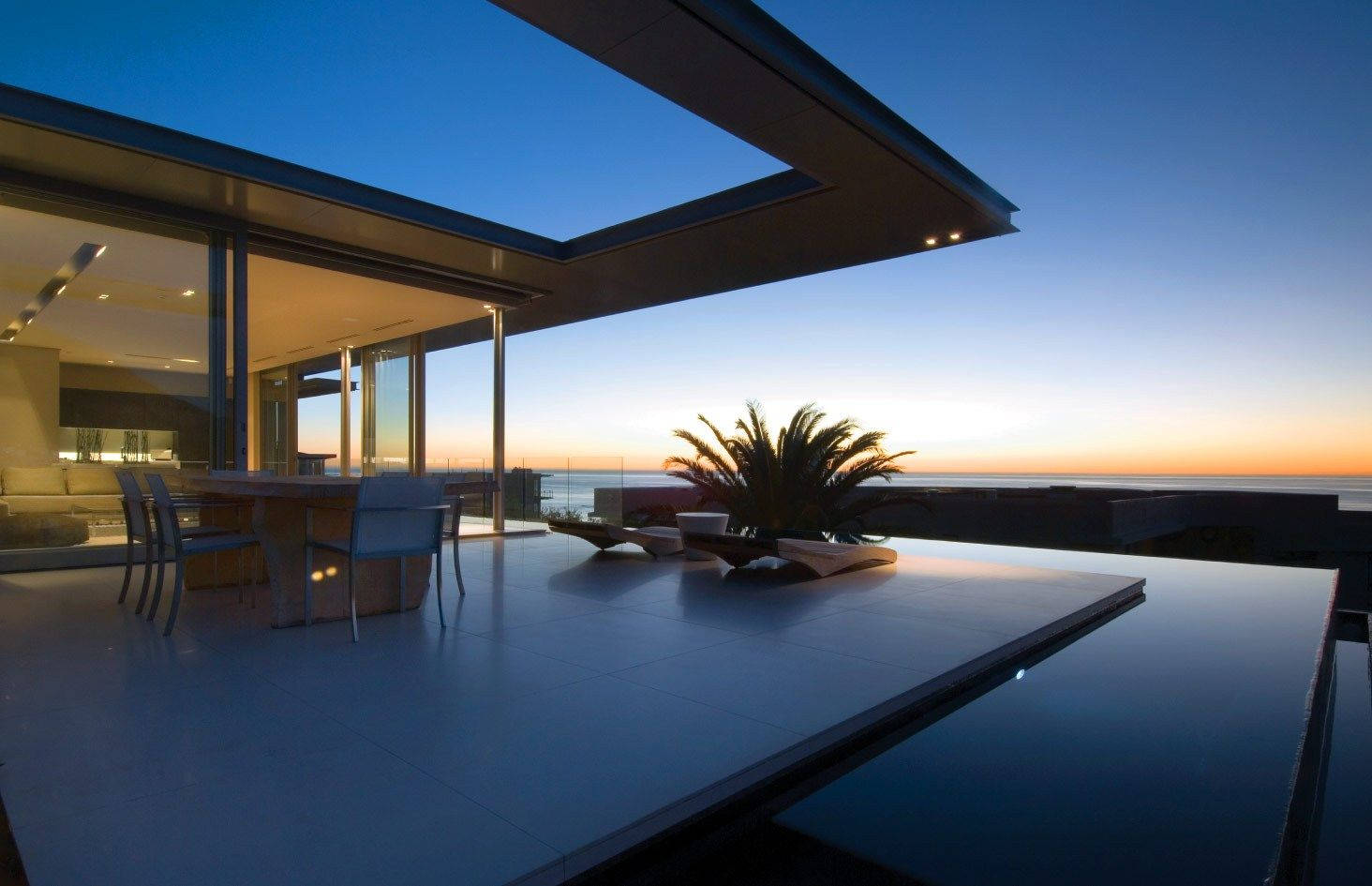 Luxury House 桌布 壁紙 — 黃昏時分的現代玻璃屋露台，設有無邊際泳池，可欣賞海景，棕櫚樹剪影映襯著暮色天空。