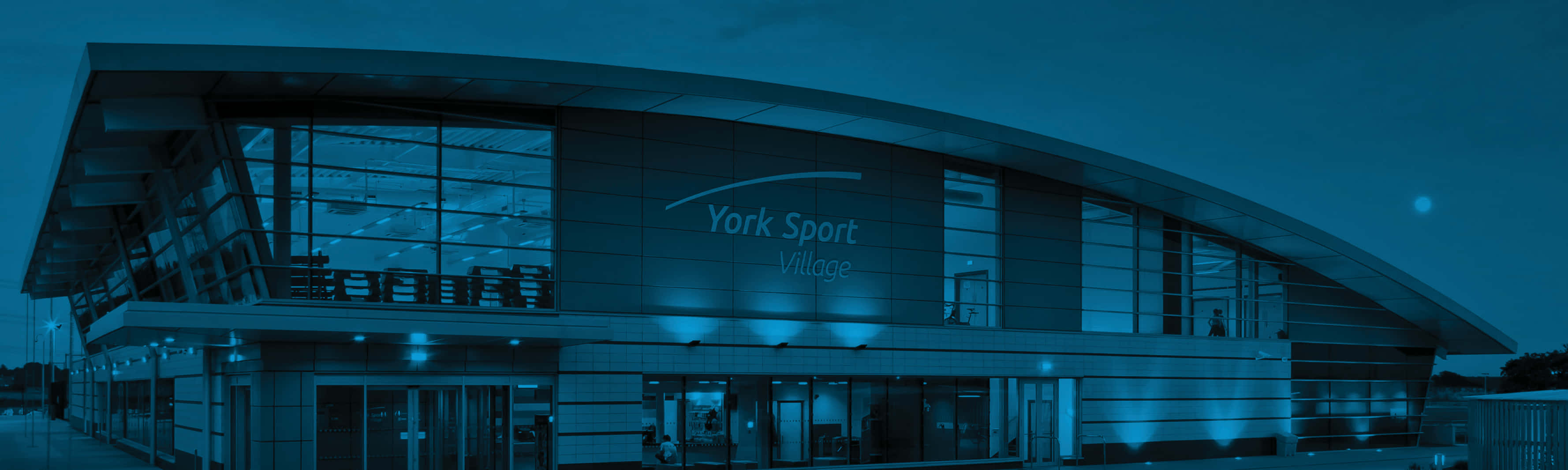 York 桌布 壁紙 — 黃昏時分的現代體育設施，藍色燈光照亮其玻璃外牆和弧形屋頂。