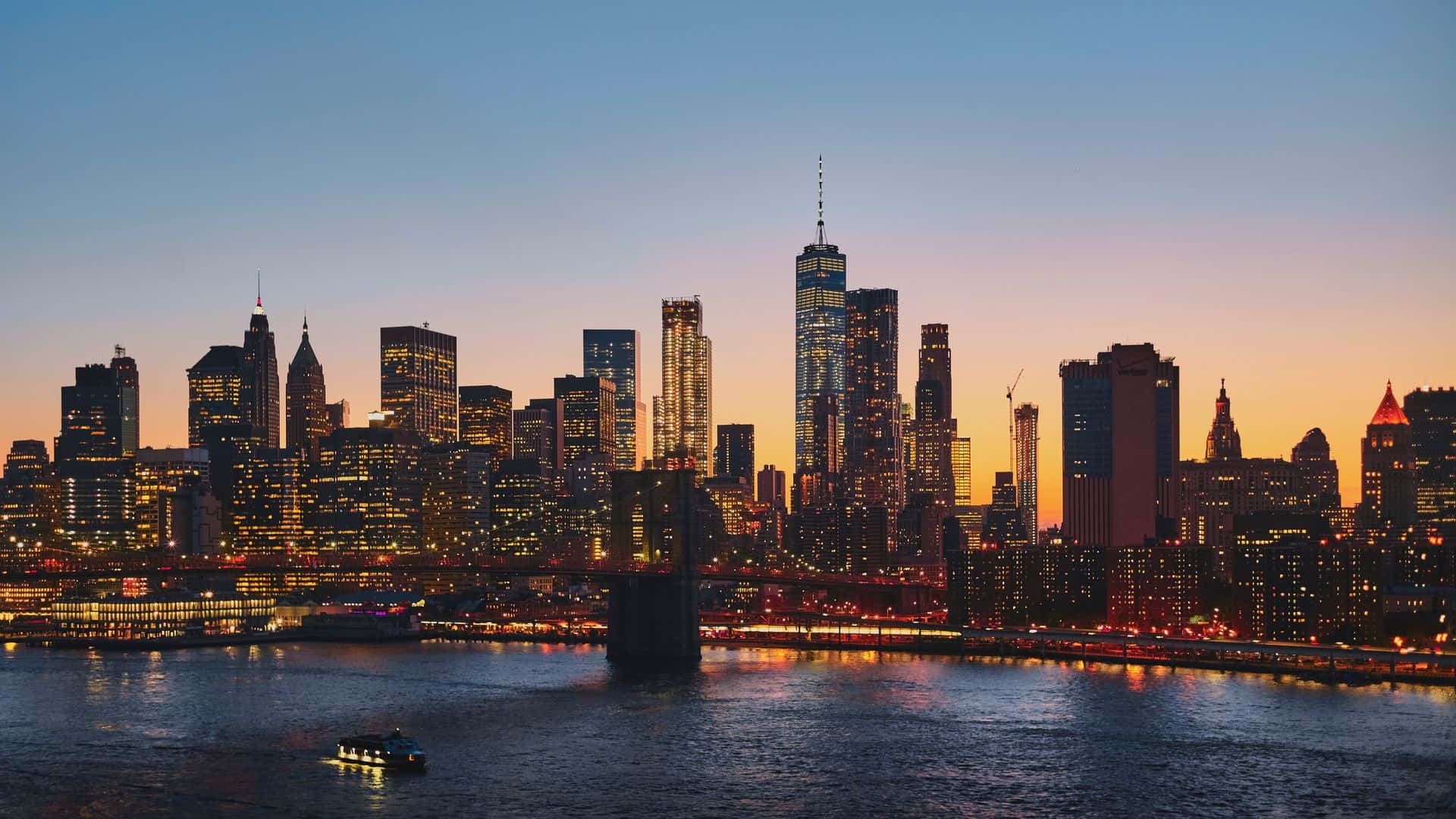 New York City At Night 桌布 壁紙 — 黃昏時分的紐約市天際線，發光的摩天大樓倒映在水面上，一艘船駛過。
