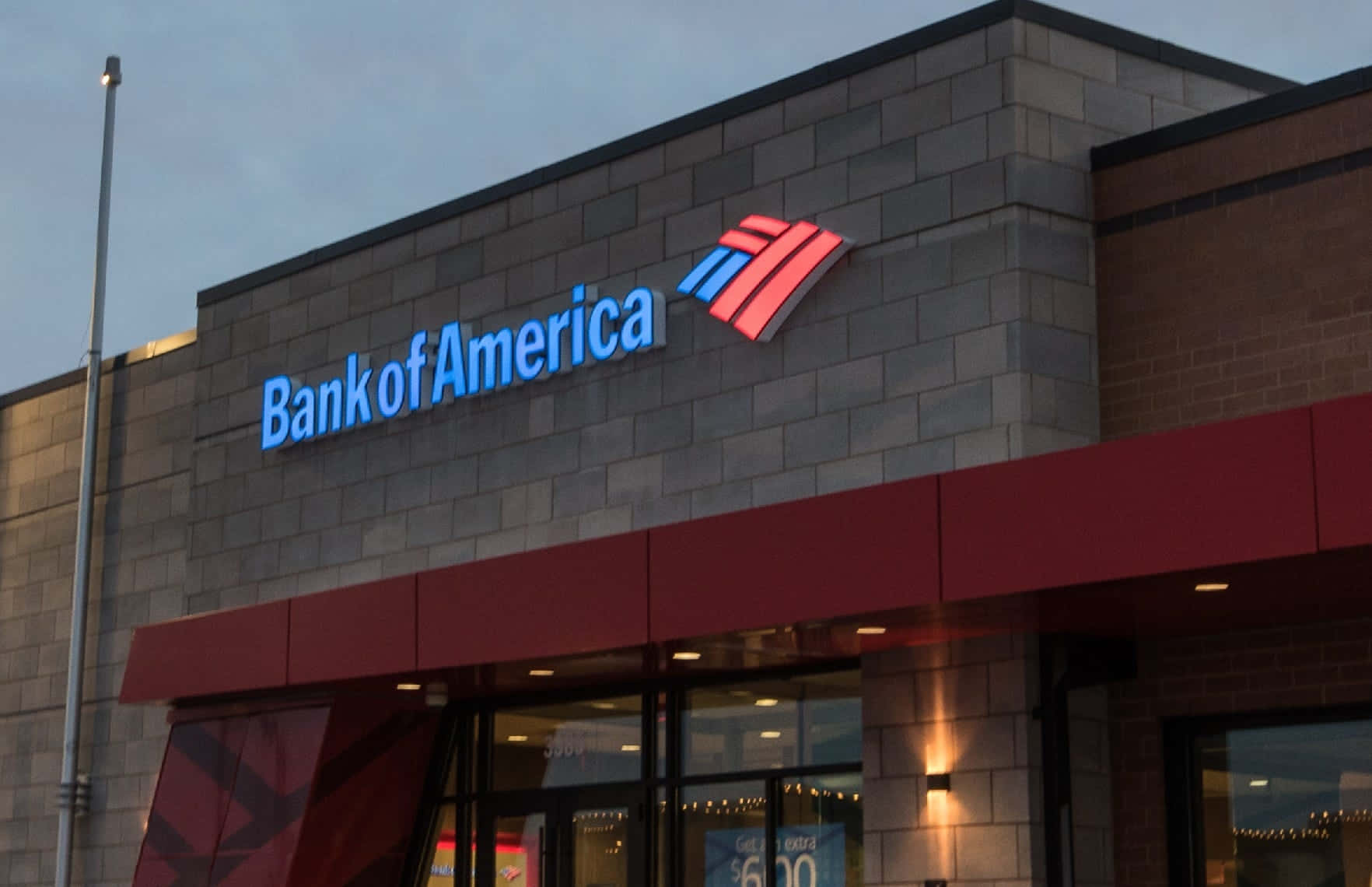 Bank Of America 桌布 壁紙 — 黃昏時分的美國銀行分行大樓，灰色磚牆外牆上閃爍著藍色和紅色的標誌。