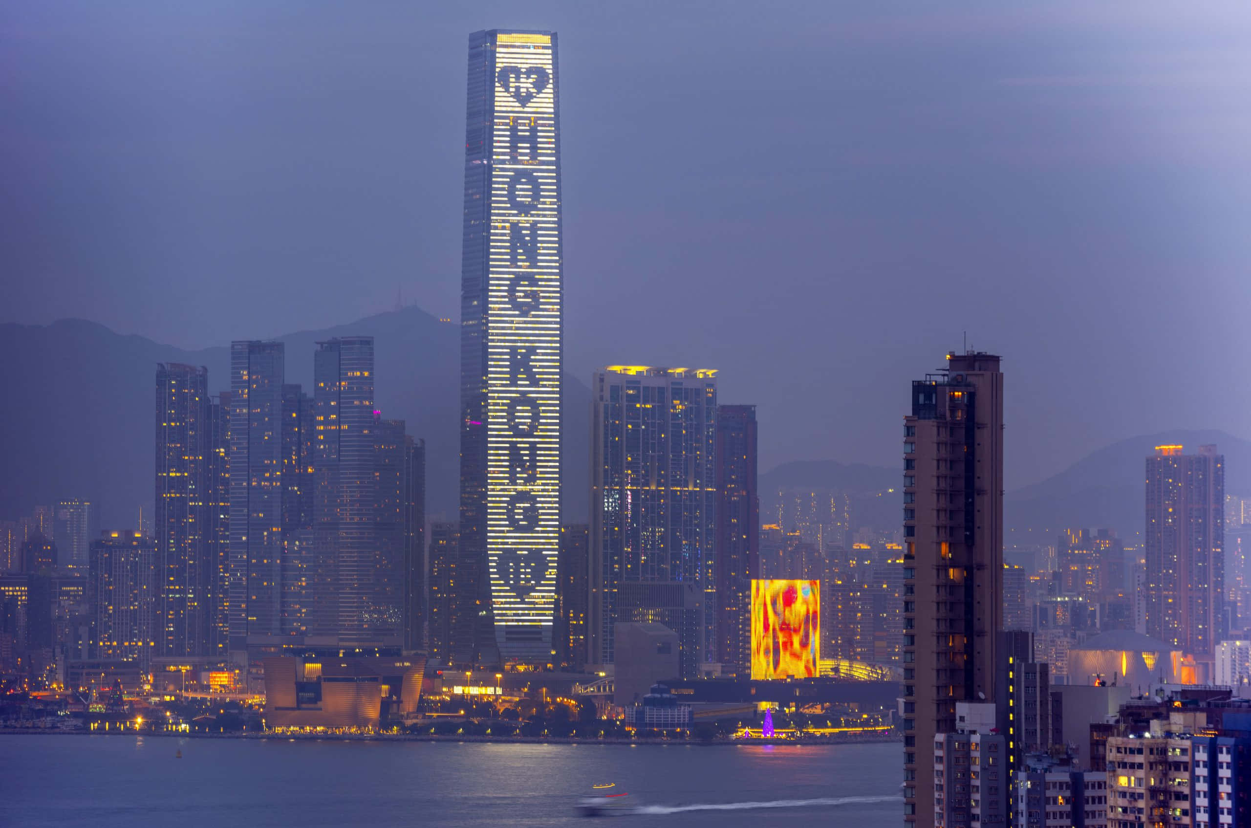 香港 桌布 壁紙 — 黃昏時分的香港天際線，一座高聳的摩天大樓在港口上方以發光字體顯示「HONG KONG」。