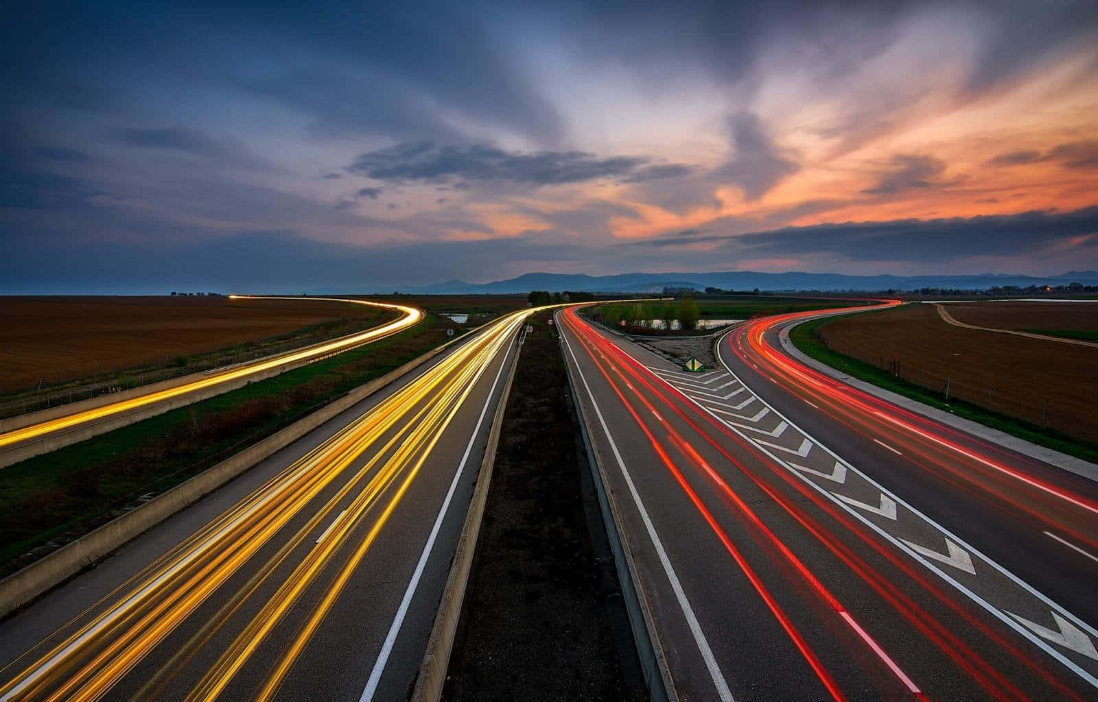 highway 桌布 壁紙 — 黃昏時分的高速公路，紅色和黃色的車燈條紋在戲劇性的天空下流過鄉村景觀。
