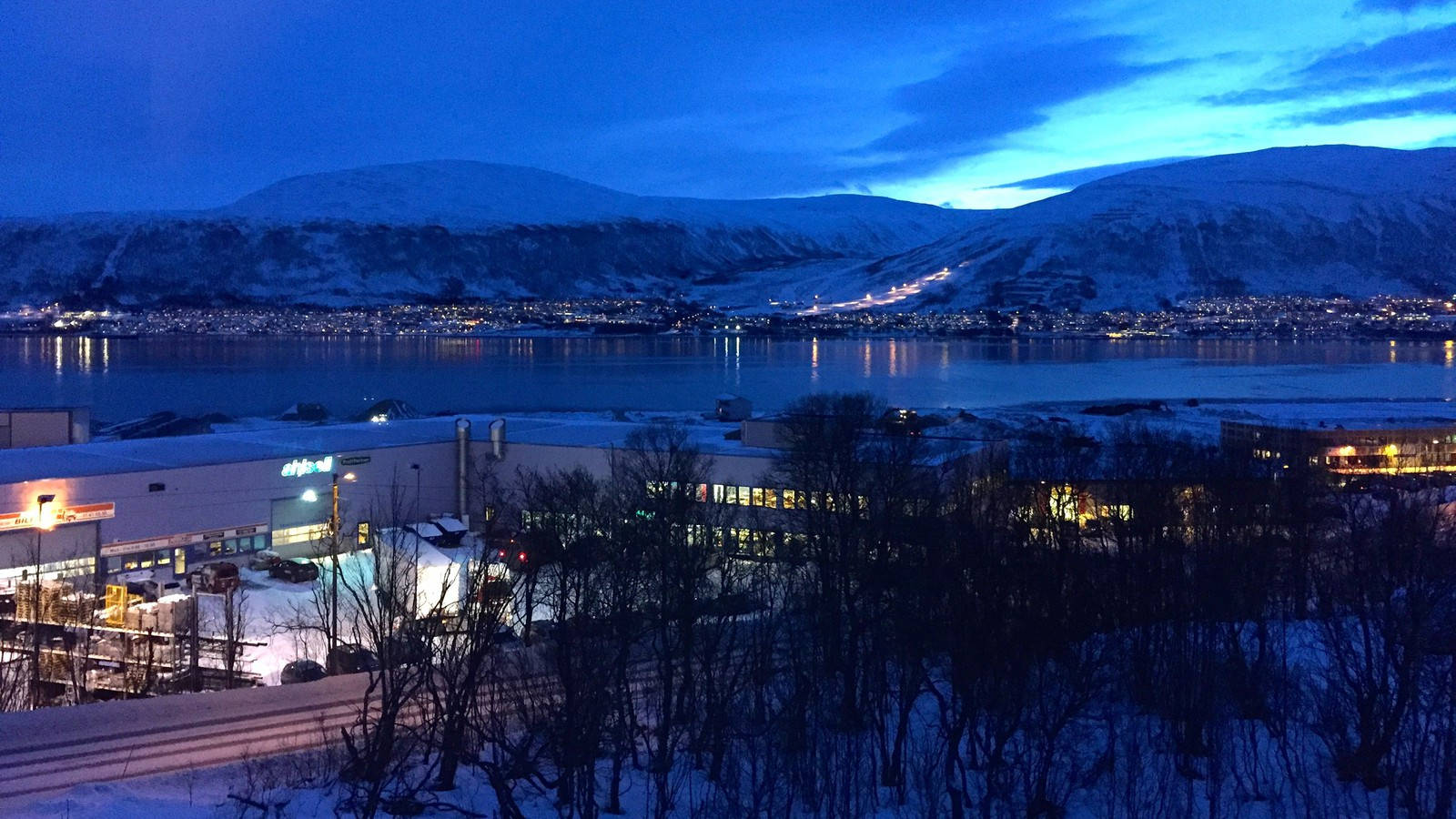 挪威 桌布 壁紙 — 黃昏時分積雪的山城，發光的燈光倒映在平靜的峽灣和前景的禿樹上。
