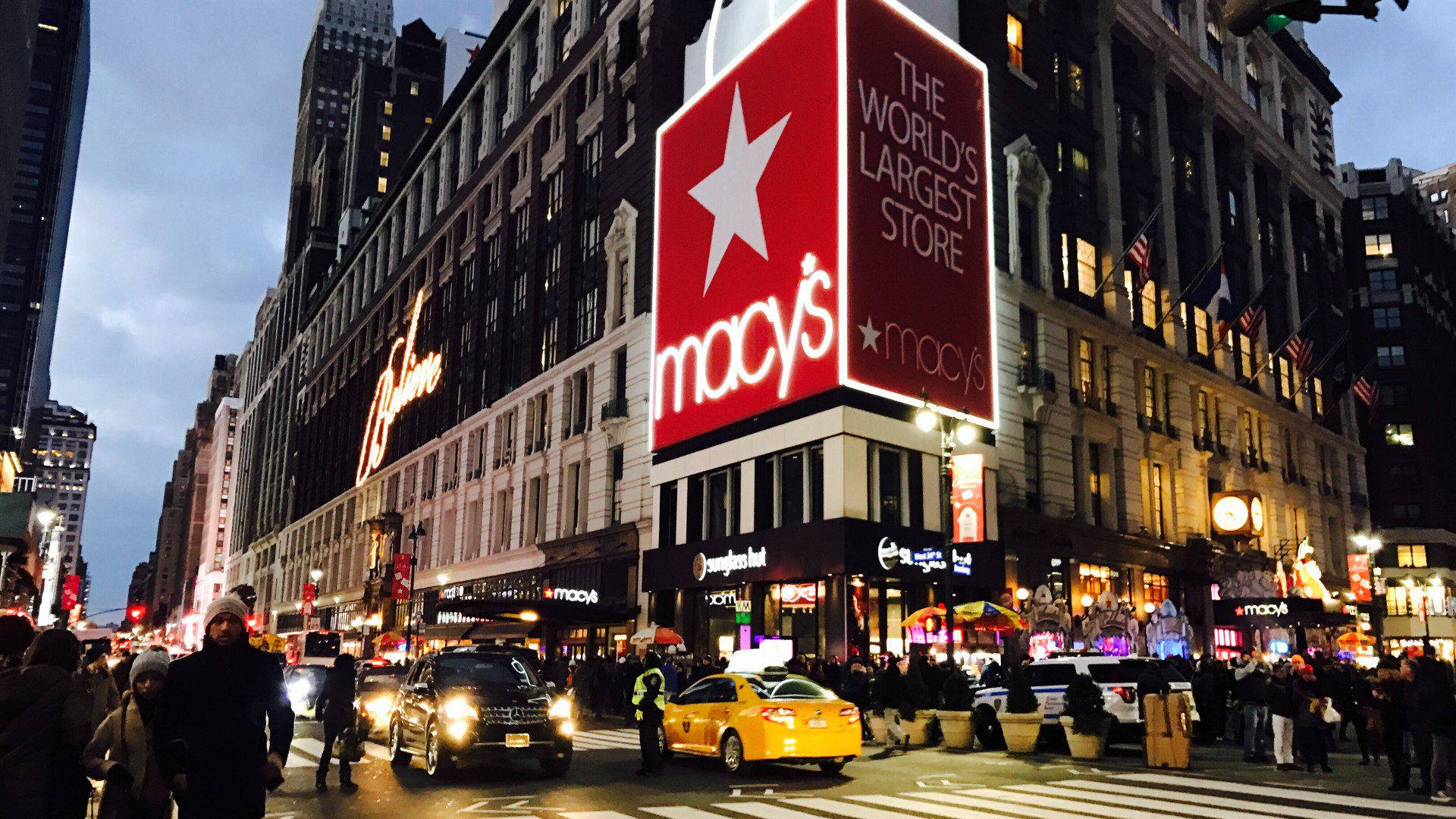 macys 桌布 壁紙 — 黃昏時分紐約市一條繁忙的街道，巨大的發光Macy's標誌聳立在行人和黃色計程車之上。