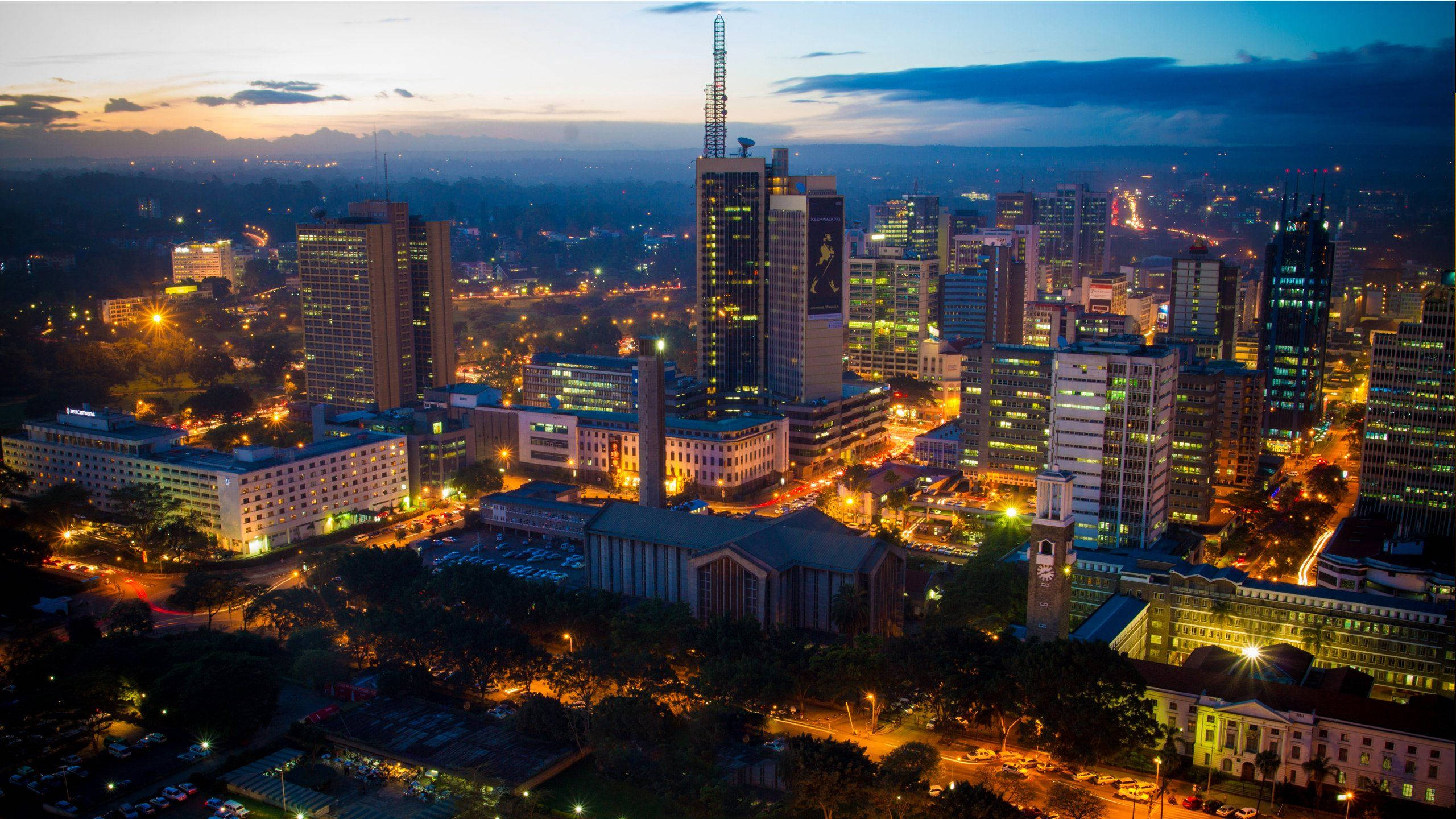 Kenya 桌布 壁紙 — 黃昏時分繁忙城市天際線的鳥瞰圖，摩天大樓燈火通明，溫暖的路燈在漸暗的藍天下閃耀。