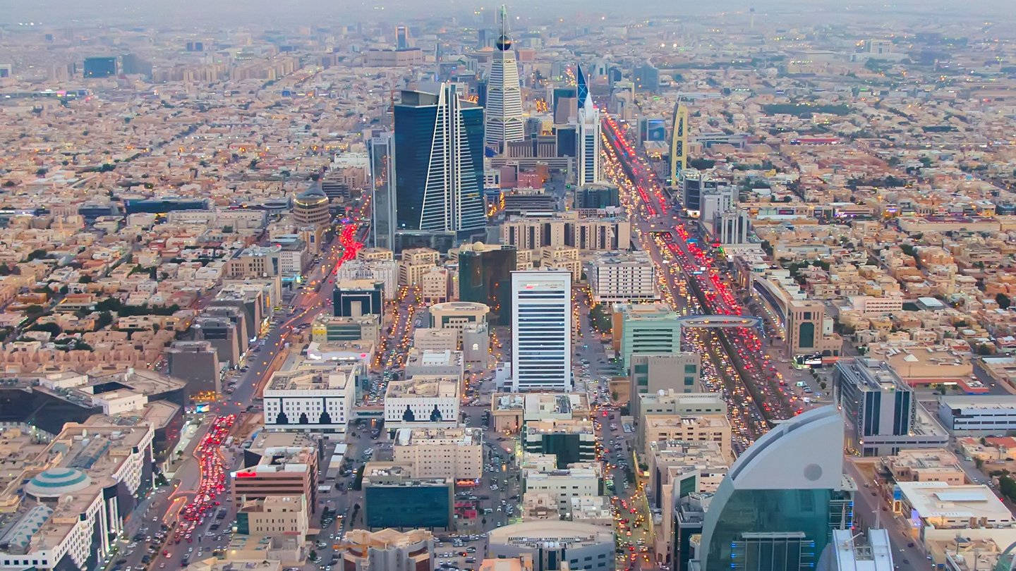 Riyadh 桌布 壁紙 — 黃昏時分繁忙城市景觀的鳥瞰圖，發光的交通和高聳的摩天大樓刺穿朦朧的天空。