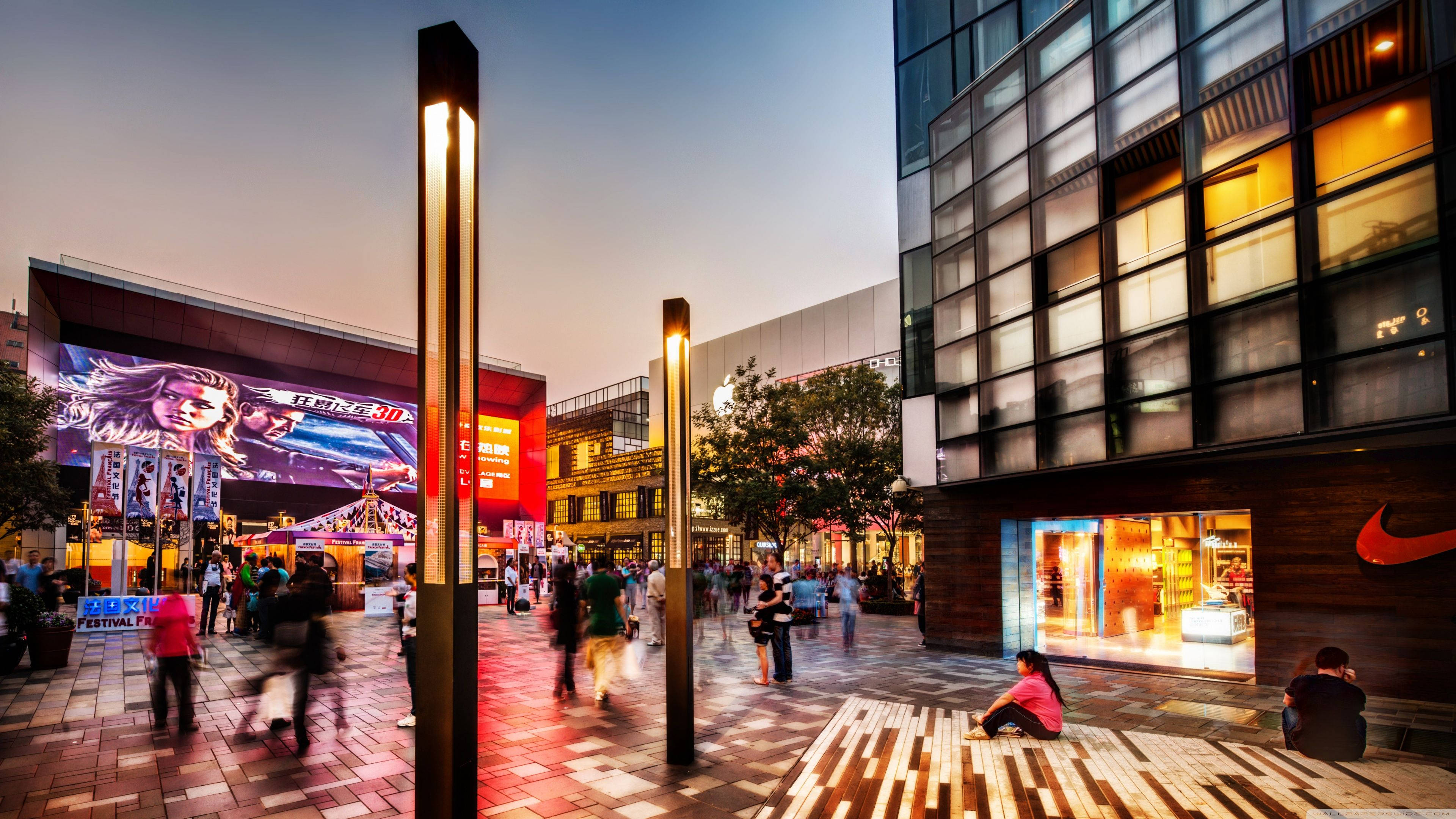 Mall 桌布 壁紙 — 黃昏時分繁忙的城市廣場，帶有發光的垂直燈光和模糊的行人，附近有Nike商店和大型數位廣告看板。