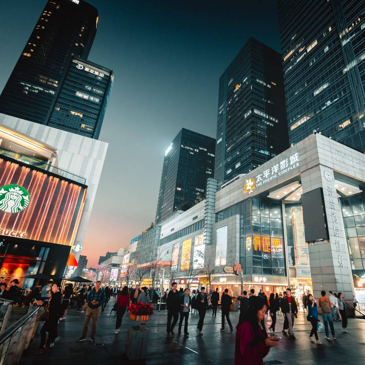 Shenzhen 桌布 壁紙 — 黃昏時分繁忙的城市廣場，高聳的玻璃摩天大樓和閃爍的商店櫥窗，行人漫步在暮色天空下。