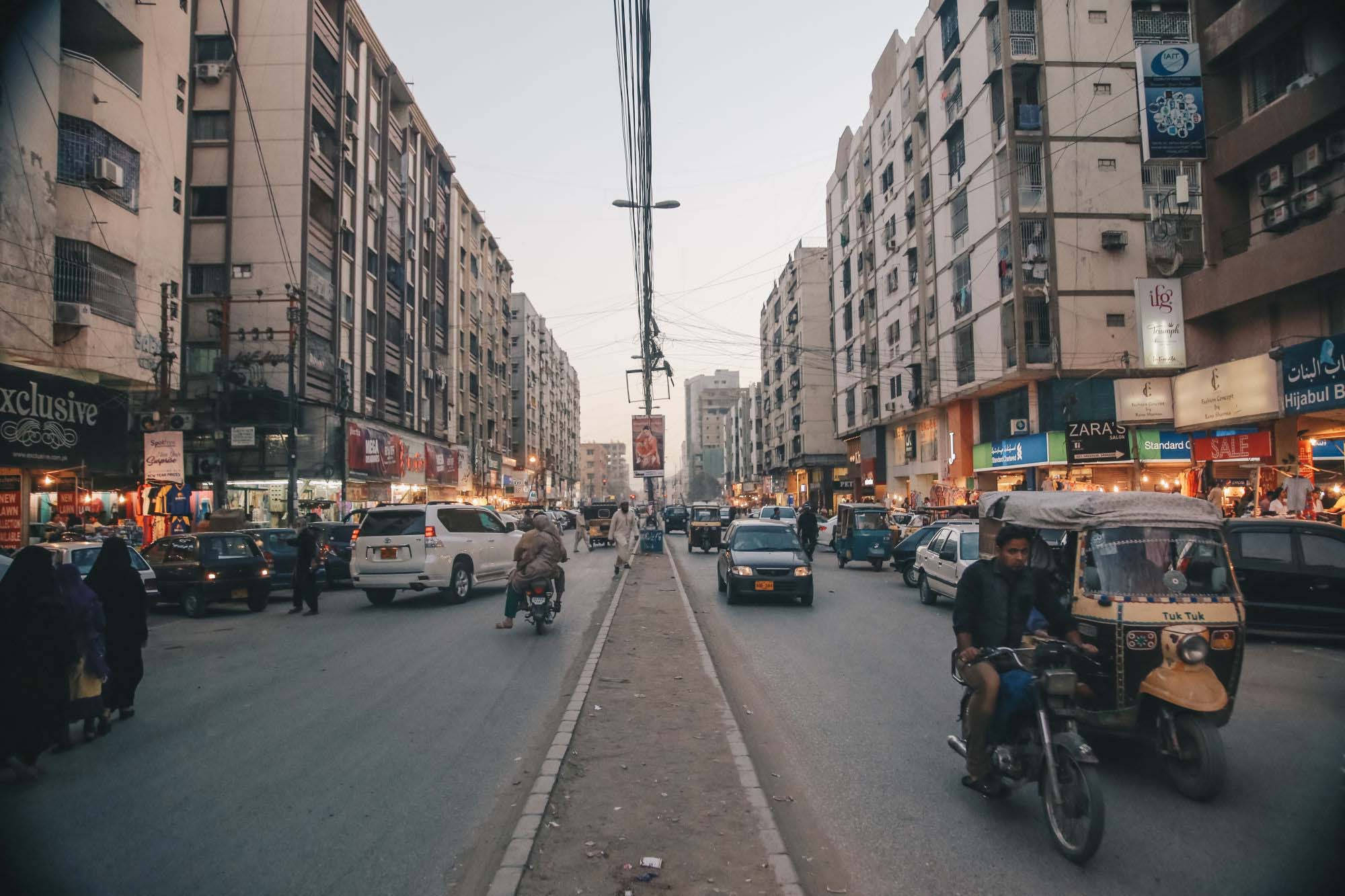 Karachi 桌布 壁紙 — 黃昏時分繁忙的城市街道，摩托車、嘟嘟車和汽車在擁擠的人行道上穿梭，兩旁是高樓大廈和發光的商店招牌。