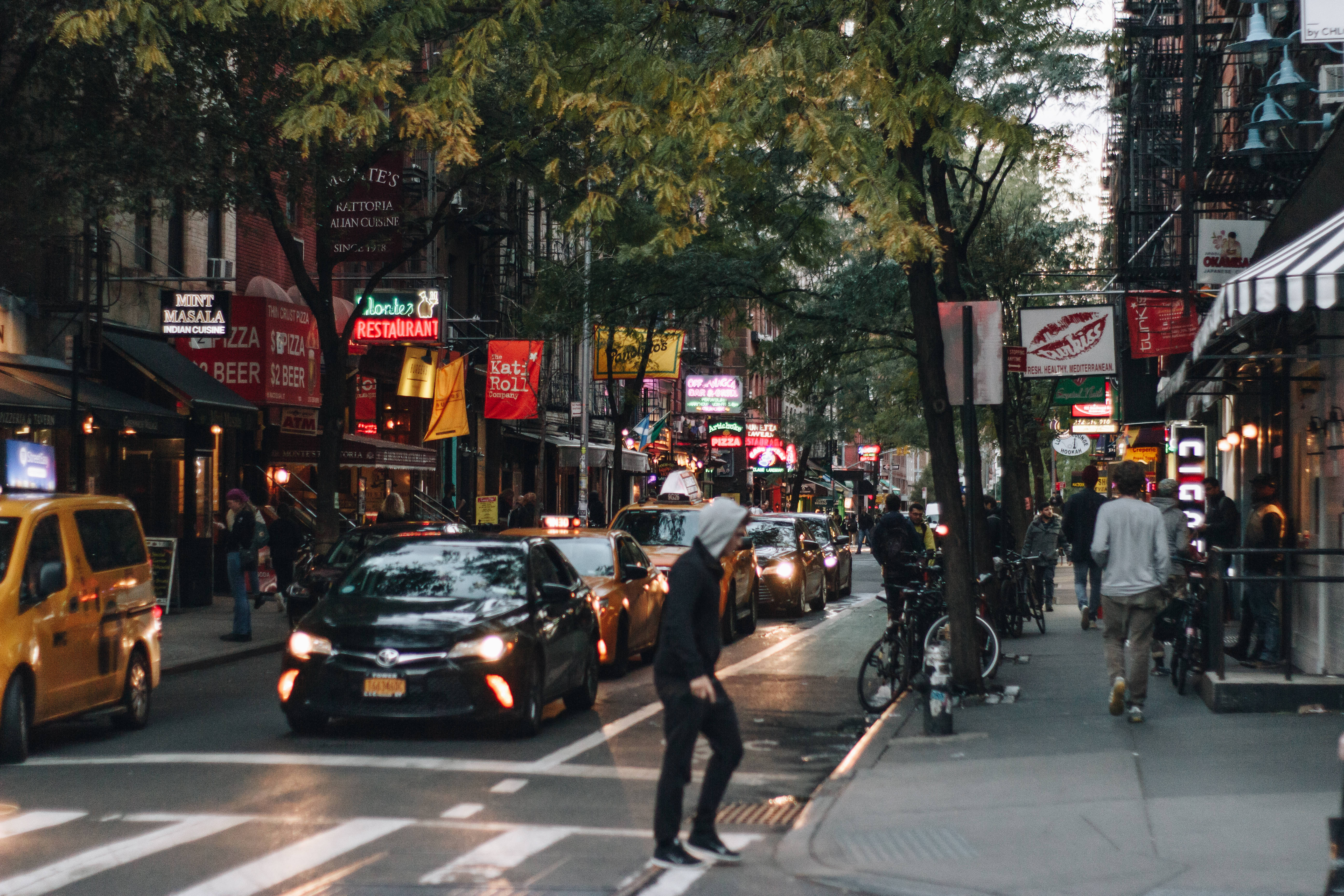 Nyc Aesthetic 桌布 壁紙 — 黃昏時分繁忙的城市街道，行人過馬路，黃色的士，以及枝葉茂密的樹下閃爍的餐廳招牌。