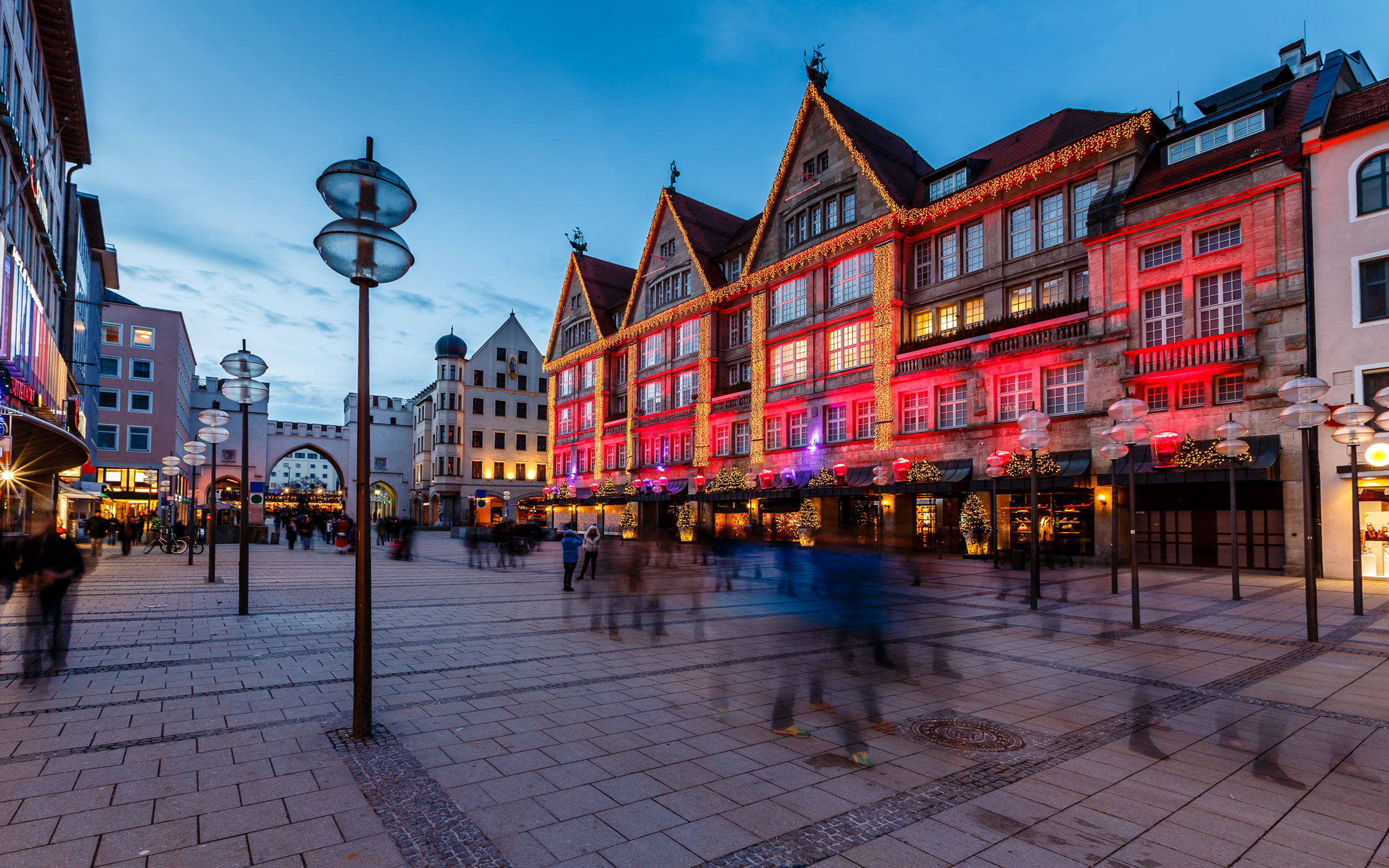 Munich 桌布 壁紙 — 黃昏時分繁忙的歐洲小鎮廣場，歷史建築在燈串下閃耀著紅色和金色光芒，模糊的行人穿過鵝卵石廣場。