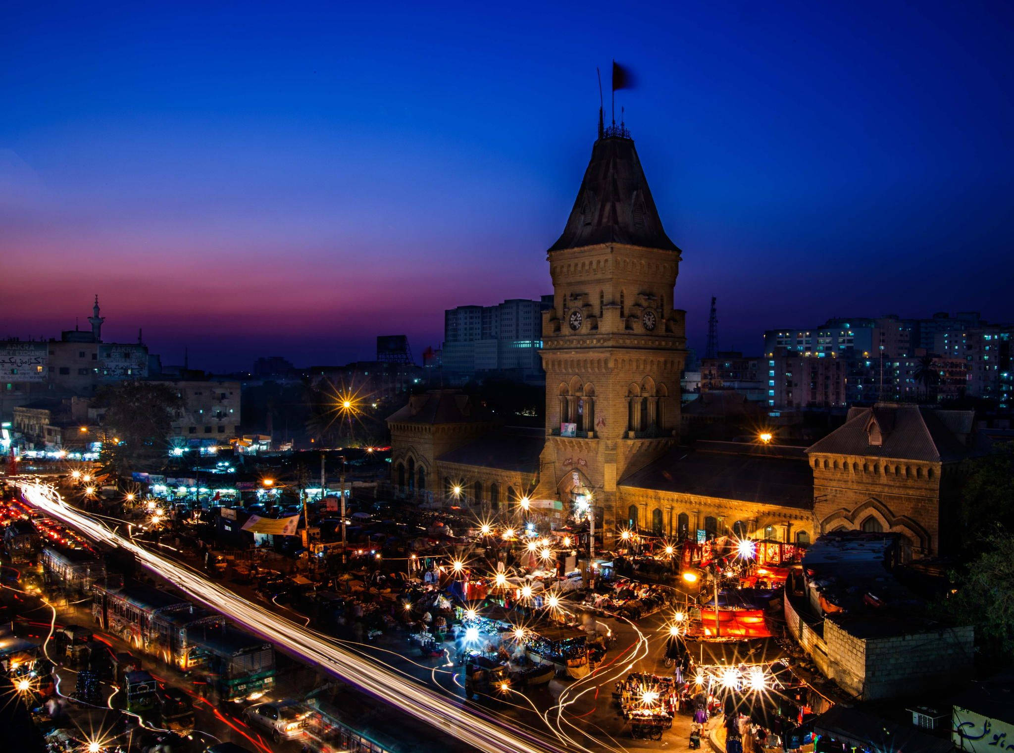 Karachi 桌布 壁紙 — 黃昏時分繁華的城市景觀，歷史悠久的鐘樓在紫藍色天空下被照亮，下方是車流燈光形成的線條。
