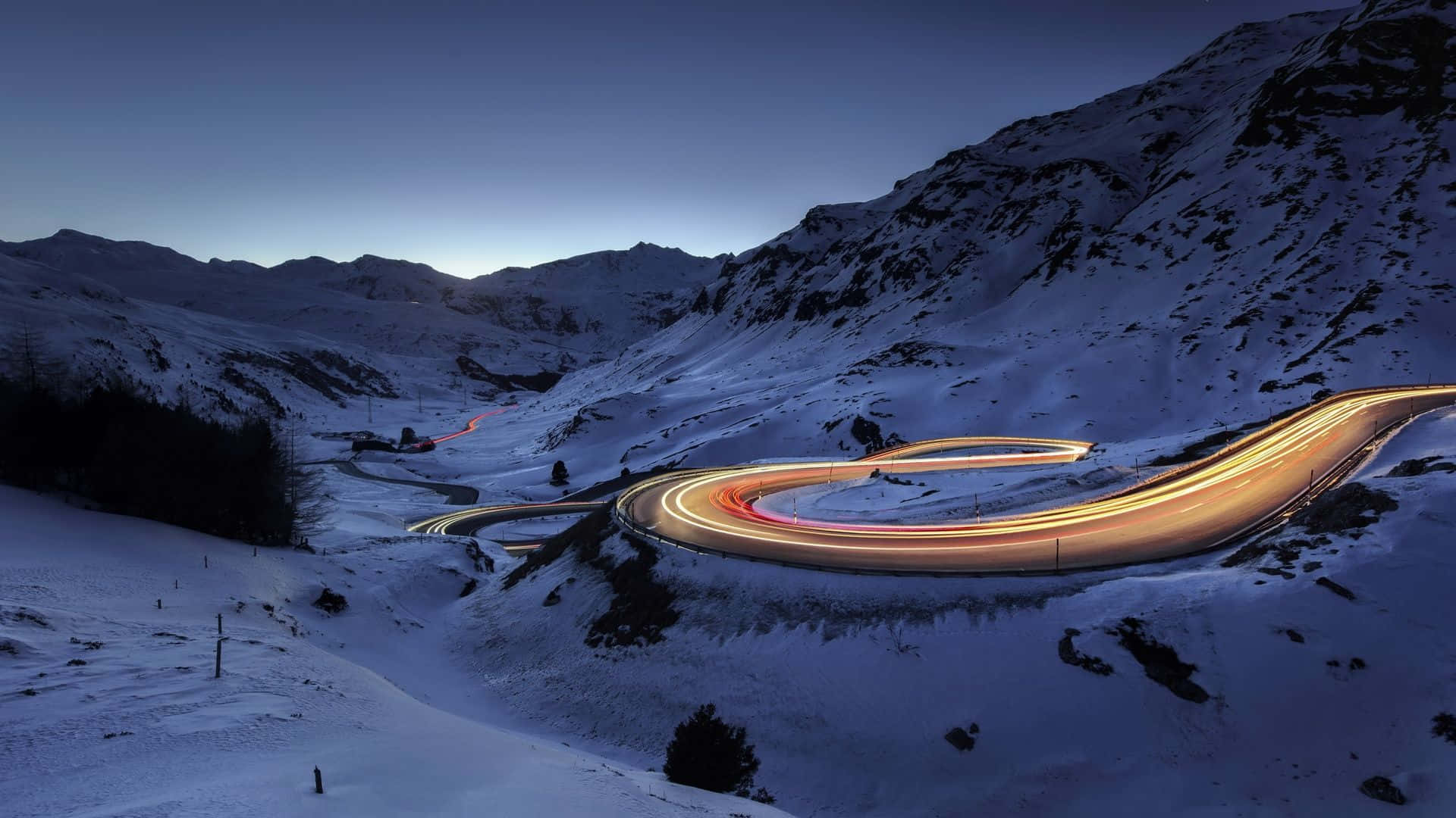 Snowy Road 桌布 壁紙 — 黃昏時分蜿蜒的山路，汽車發出的發光軌跡穿過白雪覆蓋的山峰。