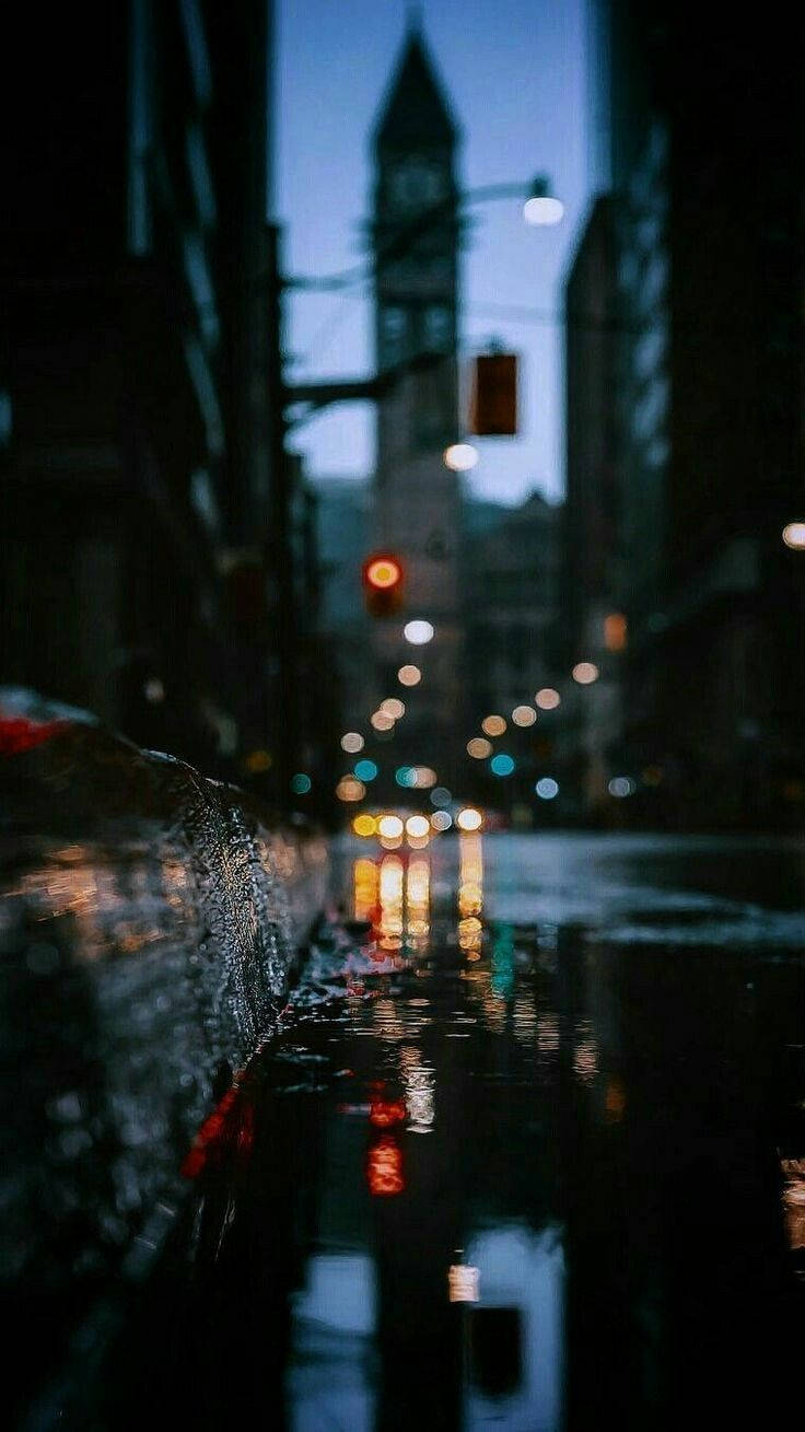 最佳高畫質 桌布 壁紙 — 黃昏時分被雨水浸濕的城市街道，模糊的路燈和遠處的鐘樓在潮濕的黑暗中柔和地發光。