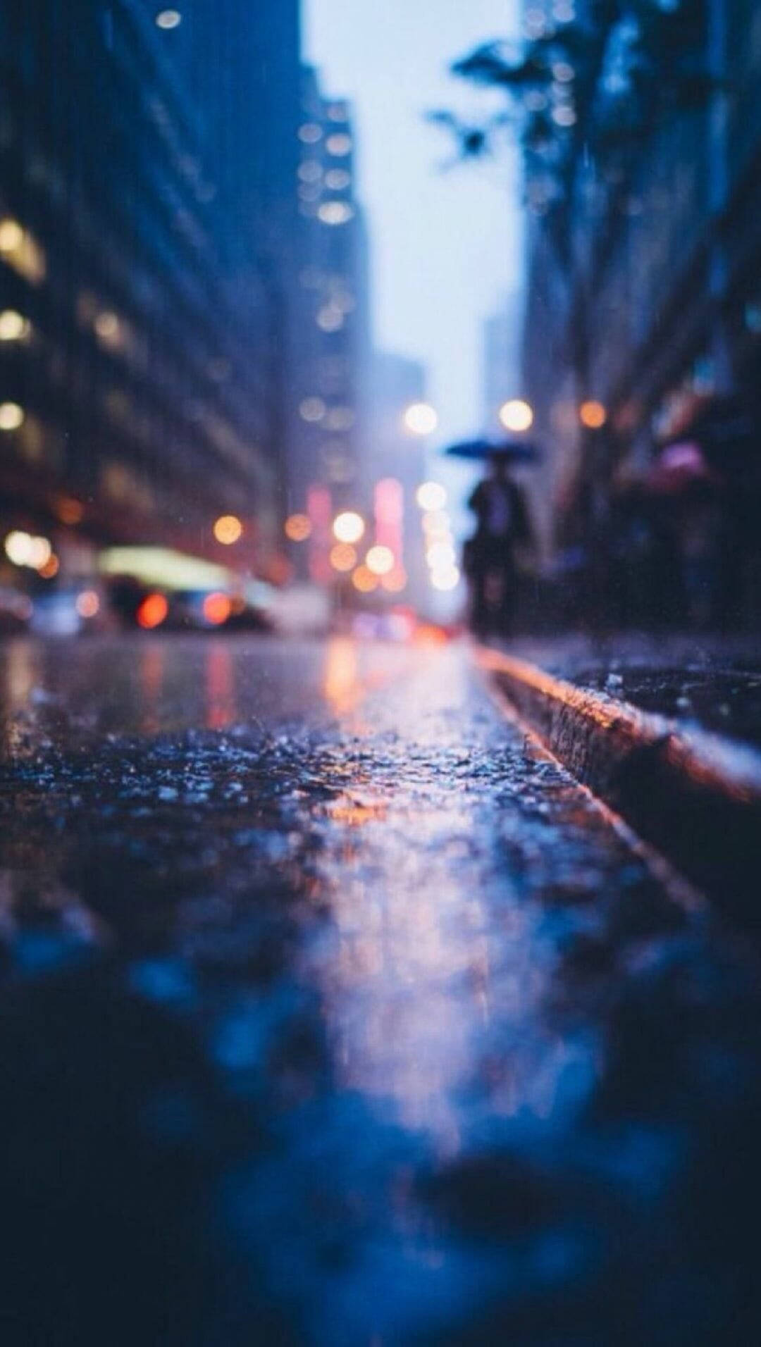 Rainy Day 桌布 壁紙 — 黃昏時分被雨水浸濕的城市街道，模糊的燈光在濕漉漉的路面上反射，一個孤獨的身影撐著傘。