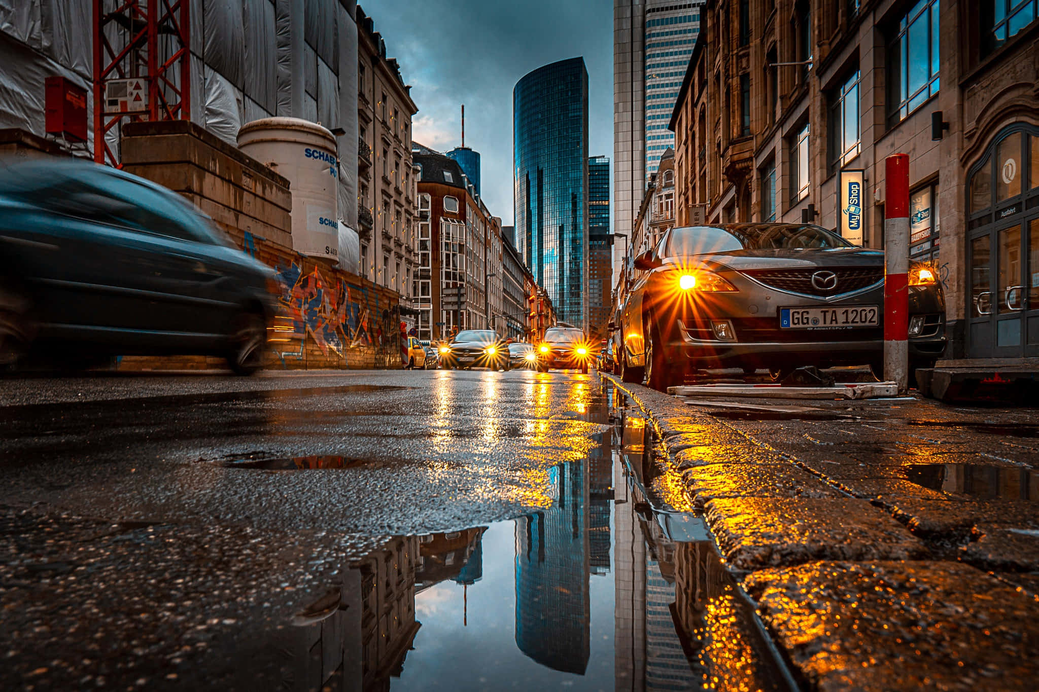 德國 桌布 壁紙 — 黃昏時分被雨水浸濕的城市街道，車頭燈在鵝卵石和現代摩天大樓之間的水坑中閃爍反射。