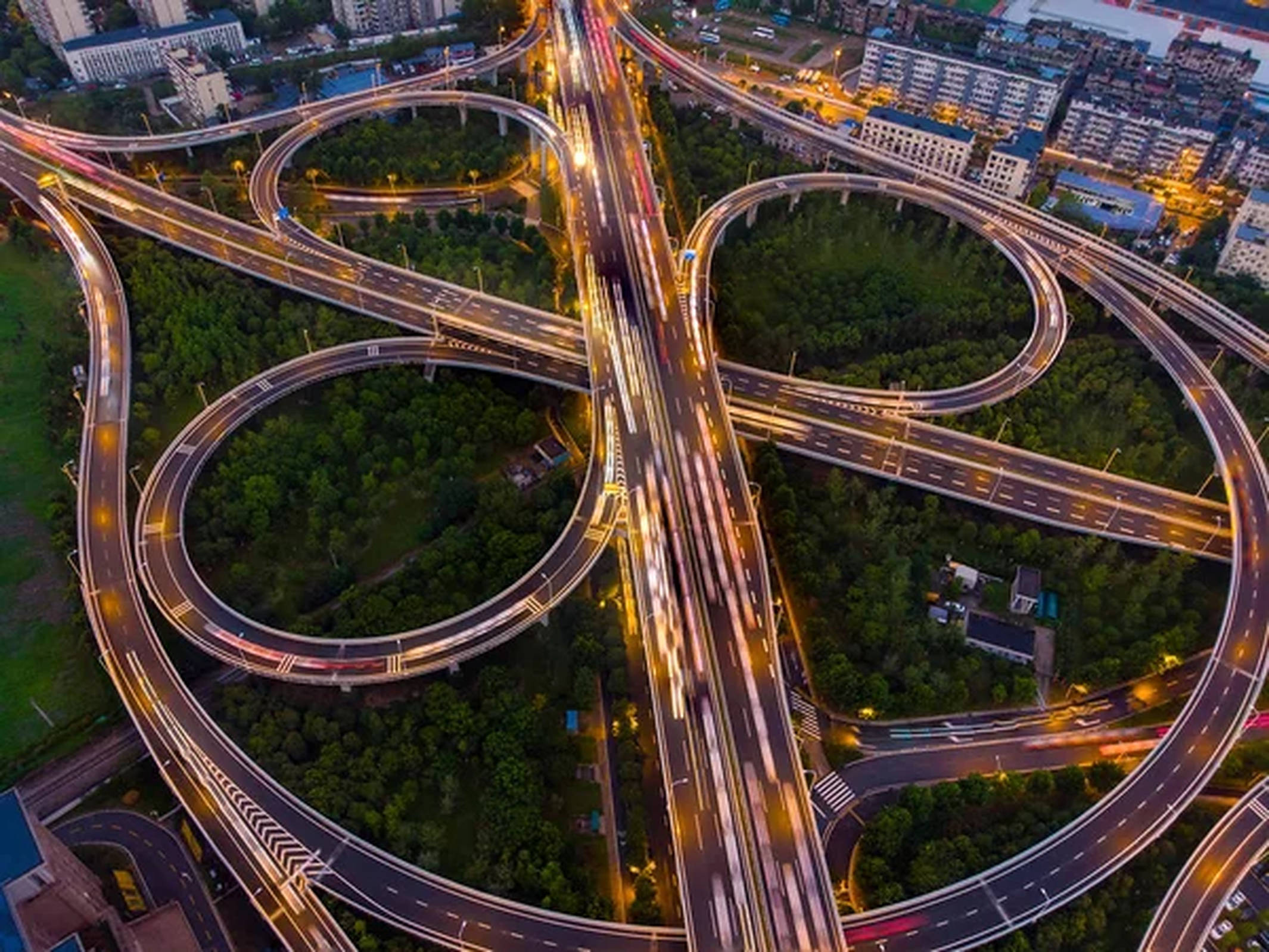 Wuhan 桌布 壁紙 — 黃昏時分複雜高速公路交匯處的鳥瞰圖，移動中的汽車發出閃爍的光軌，蜿蜒穿過茂密的綠植。