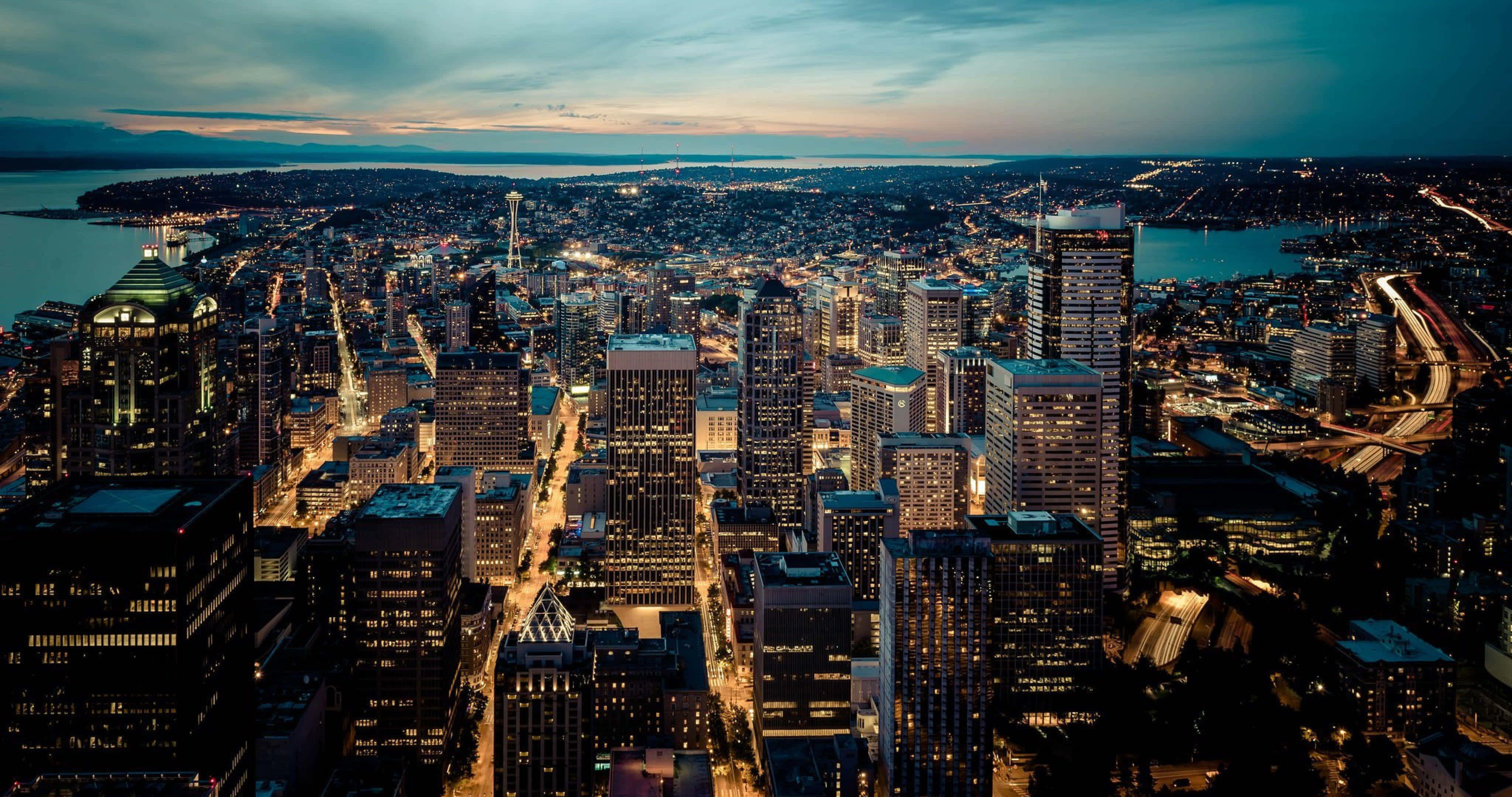 Seattle At Night 桌布 壁紙 — 黃昏時分西雅圖天際線的鳥瞰圖，閃爍的摩天大樓和海濱燈光倒映在水面上。