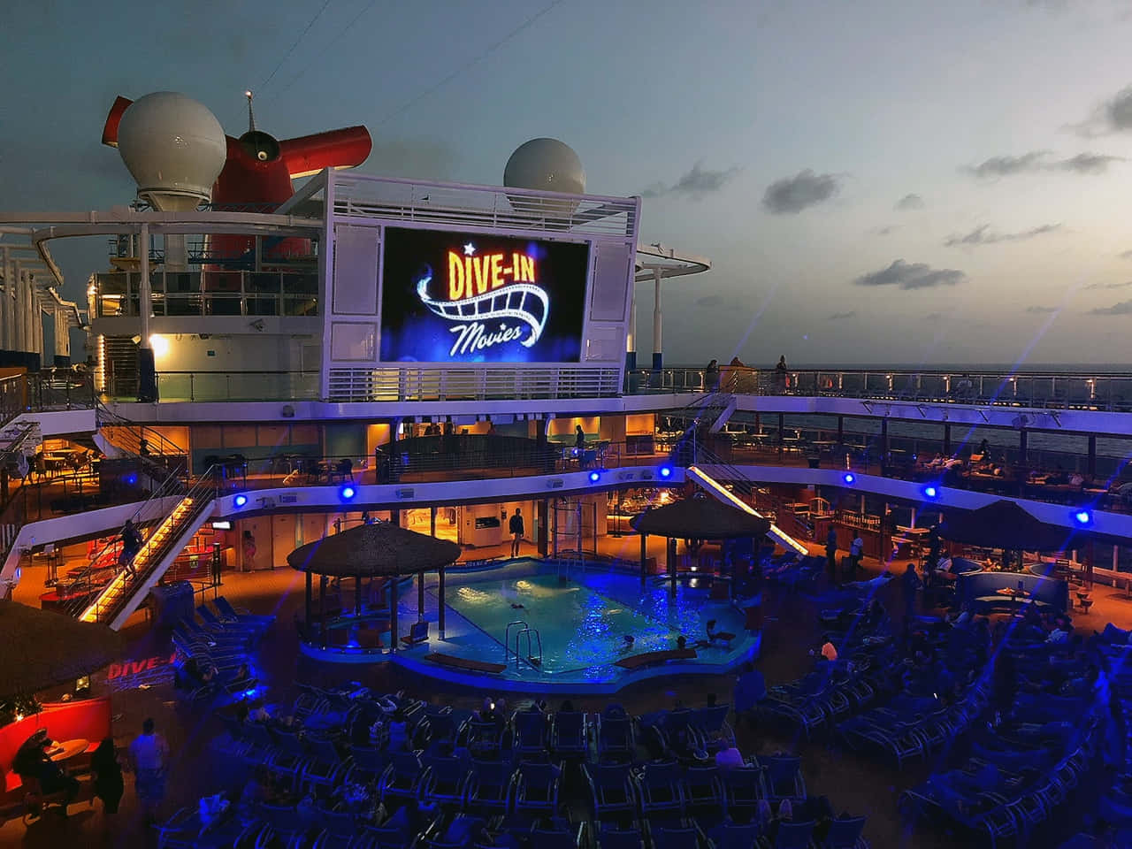 Carnival Conquest 桌布 壁紙 — 黃昏時分郵輪上的戶外電影院，發光的螢幕位於泳池上方，周圍環繞著躺椅。