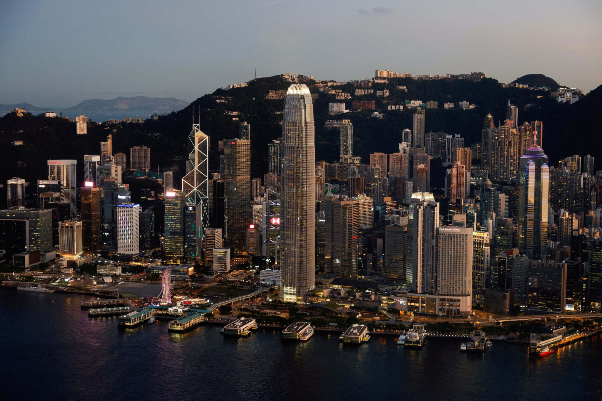香港 桌布 壁紙 — 黃昏時分香港天際線的鳥瞰圖，燈火通明的摩天大樓倒映在港灣水面和背景的山丘上。