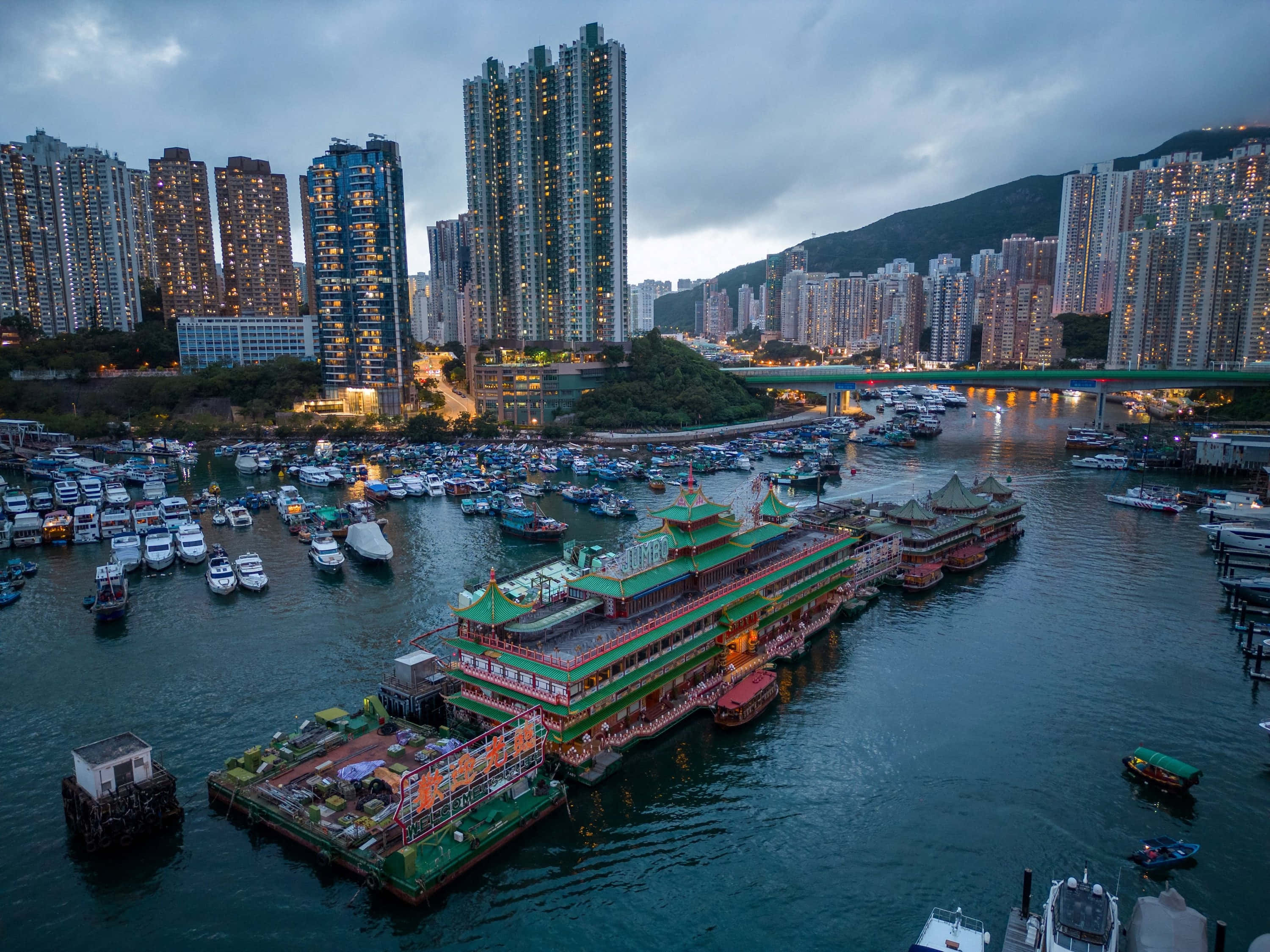 香港 桌布 壁紙 — 黃昏時分香港海港的鳥瞰圖，燈火通明的摩天大樓和傳統的綠色屋頂渡輪停泊在擁擠的水域中。