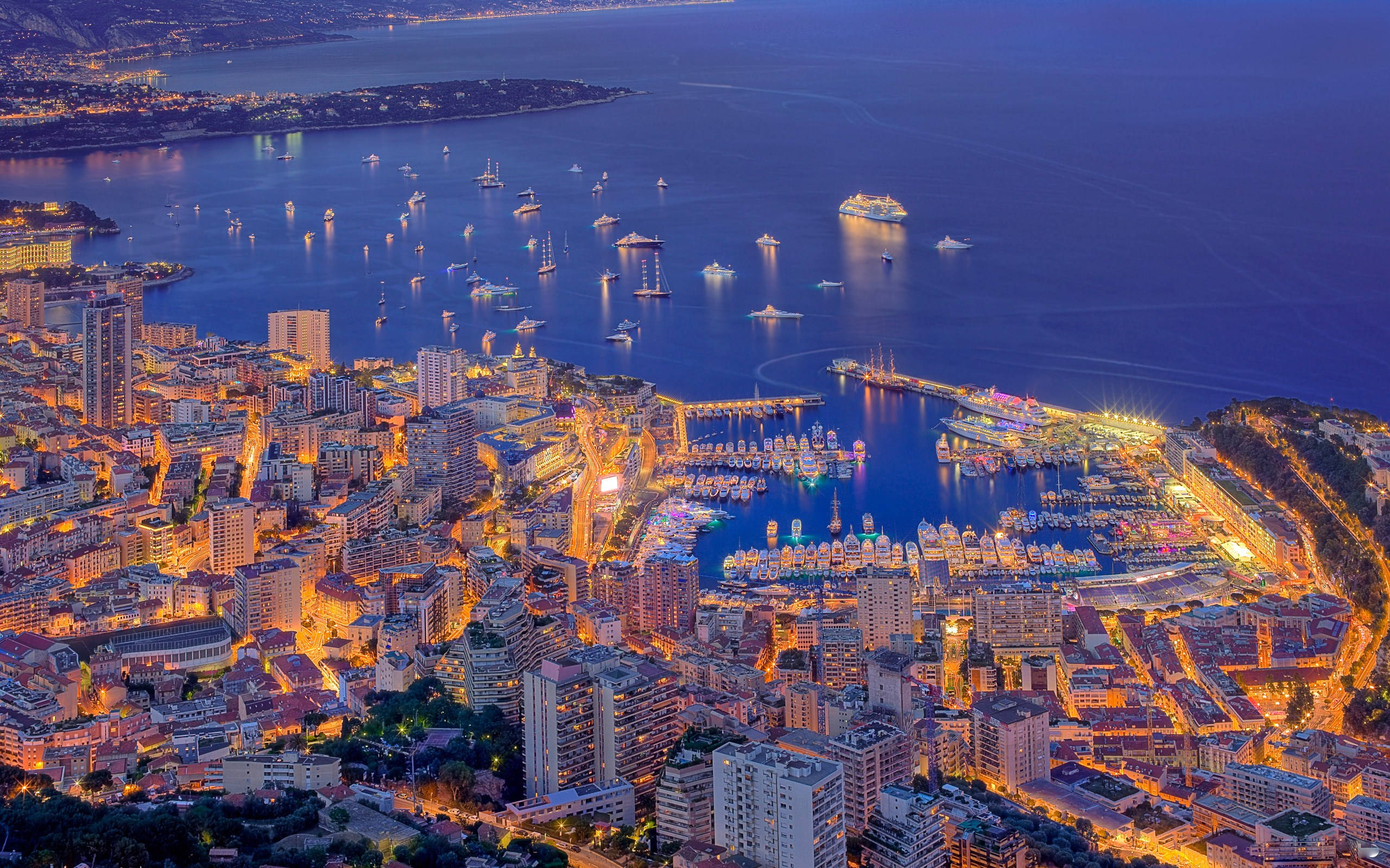 Monaco 桌布 壁紙 — 黃昏時摩納哥的鳥瞰圖，發光的城市燈光反射在停滿遊艇和郵輪的港口上。