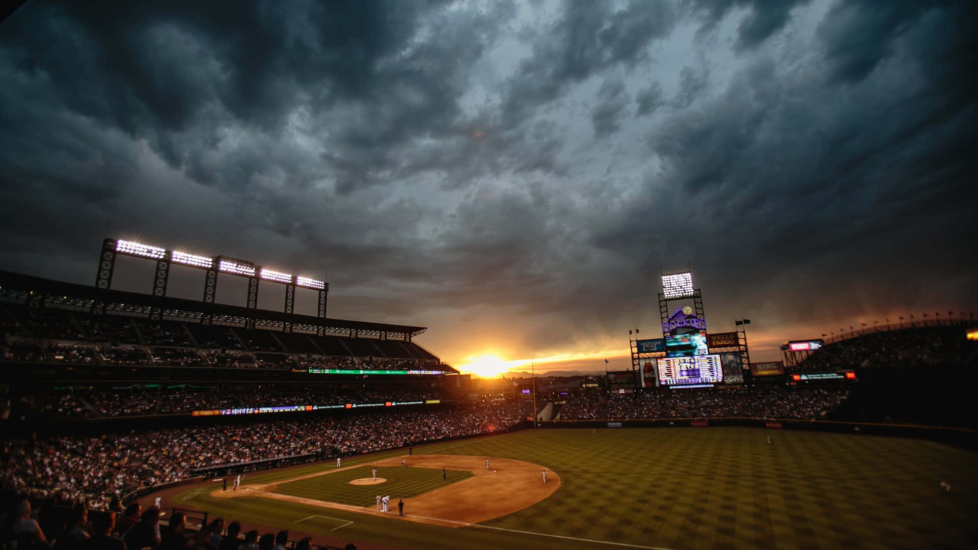 MLB Baseball League 桌布 壁紙 — 黃昏時的棒球場，戲劇性的暴風雨天空和發光的記分板照亮球場。