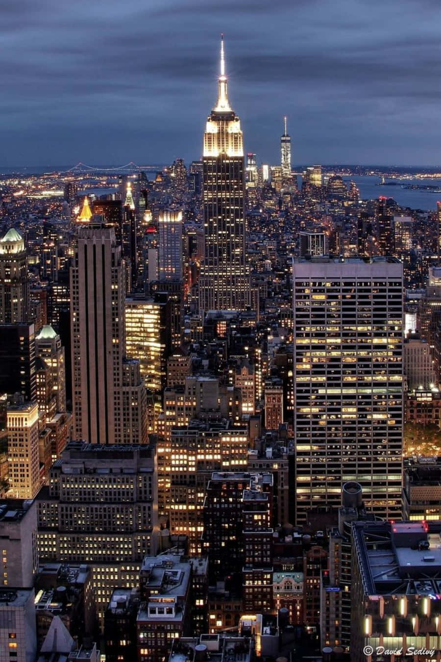 Nyc Aesthetic 桌布 壁紙 — 黃昏時紐約市天際線的鳥瞰圖，帝國大廈在黑暗的雲層中閃耀，城市燈光在下方閃爍。