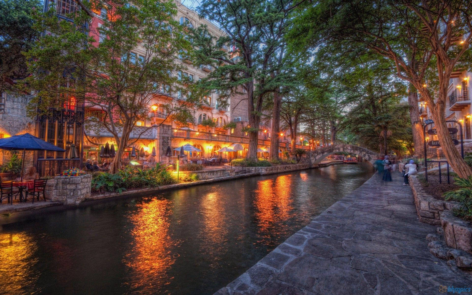 San Antonio 桌布 壁紙 — 黃昏時迷人的運河，溫暖的燈光倒映在水面上，周圍環繞著樹木和石頭步道。