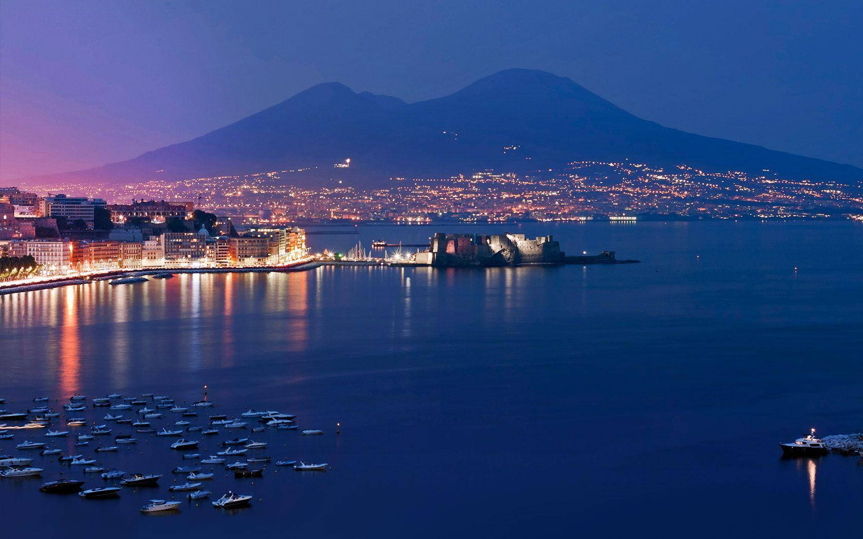 Naples 桌布 壁紙 — 黃昏海岸城市景觀，平靜水面上倒映著閃爍燈光，背景中雄偉的山巒若隱若現。