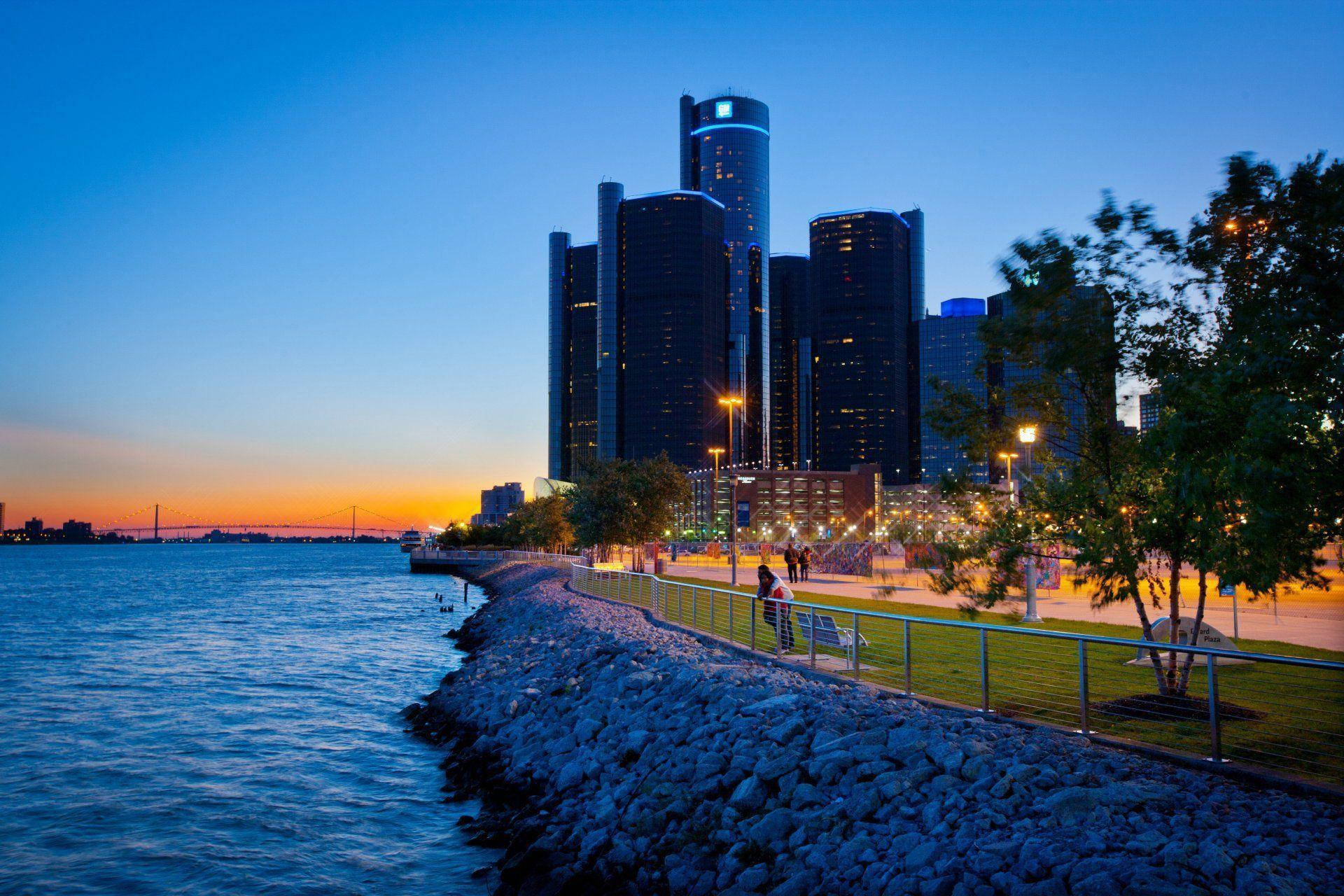 detroit 桌布 壁紙 — 黃昏海濱場景，燈火通明的摩天大樓倒映在平靜的水面上，人們沿著石堤漫步。