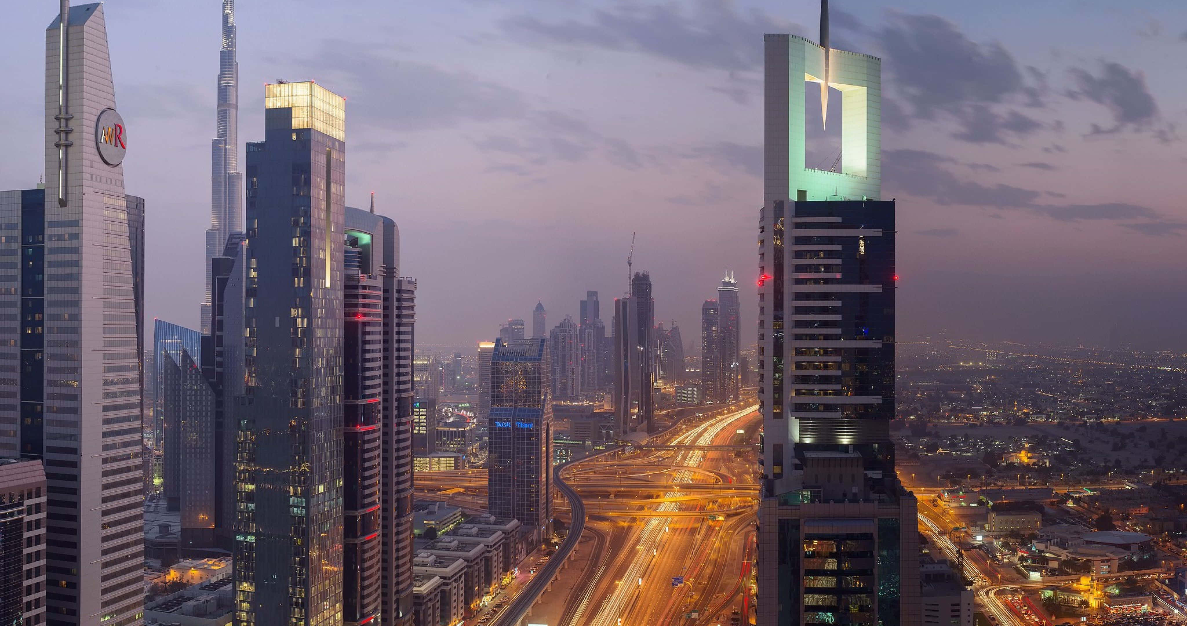 Uae 桌布 壁紙 — 黃昏照亮杜拜的天際線，摩天大樓閃閃發光，高速公路燈光在紫色天空下劃出線條。