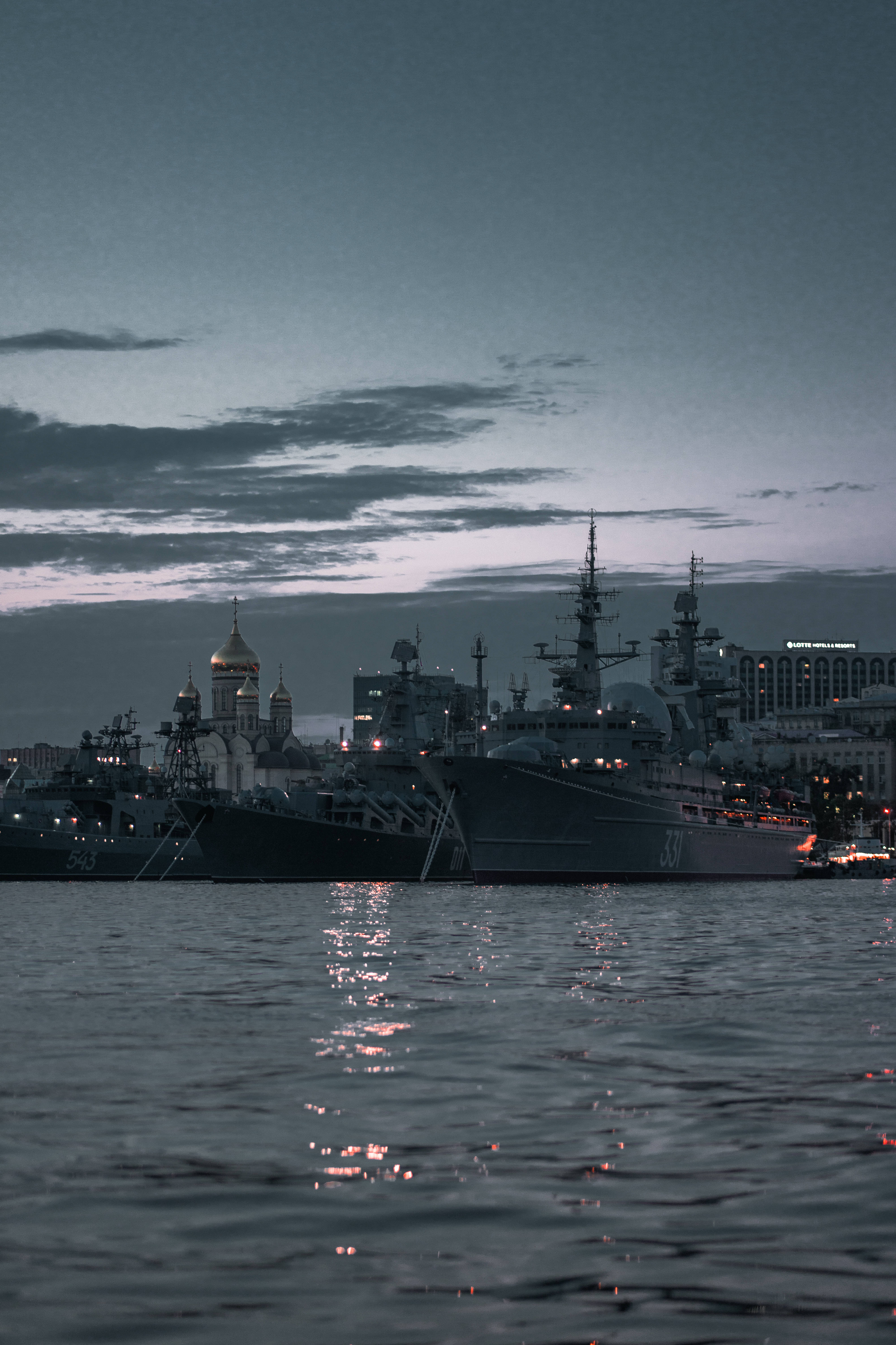 Warship 桌布 壁紙 — 黃昏降臨在一個港口，軍艦停泊在一個擁有金色圓頂教堂和水面上閃爍燈光的城市景觀旁。
