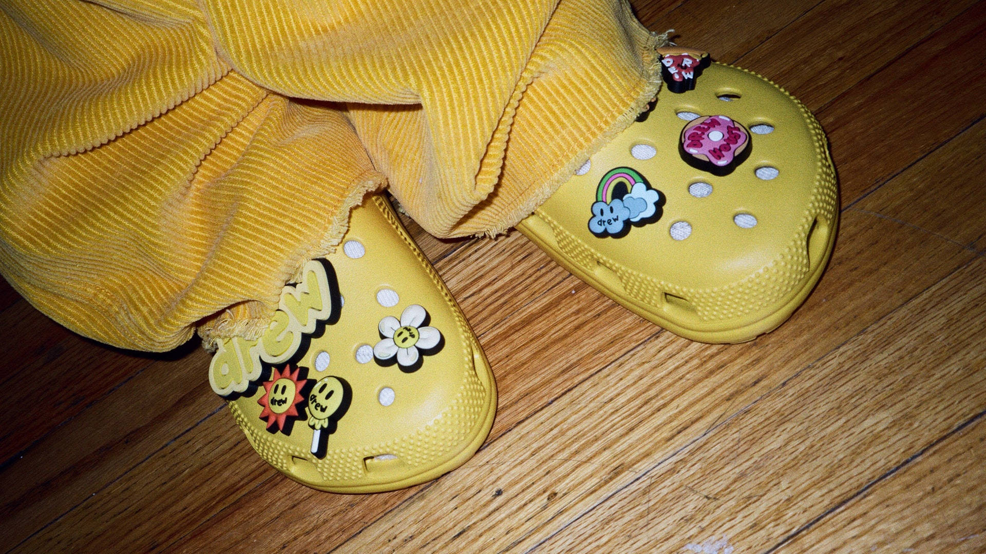 Crocs 桌布 壁紙 — 黃色Crocs鞋綴有彩色飾物，放在木地板上，下方是黃色燈芯絨褲。