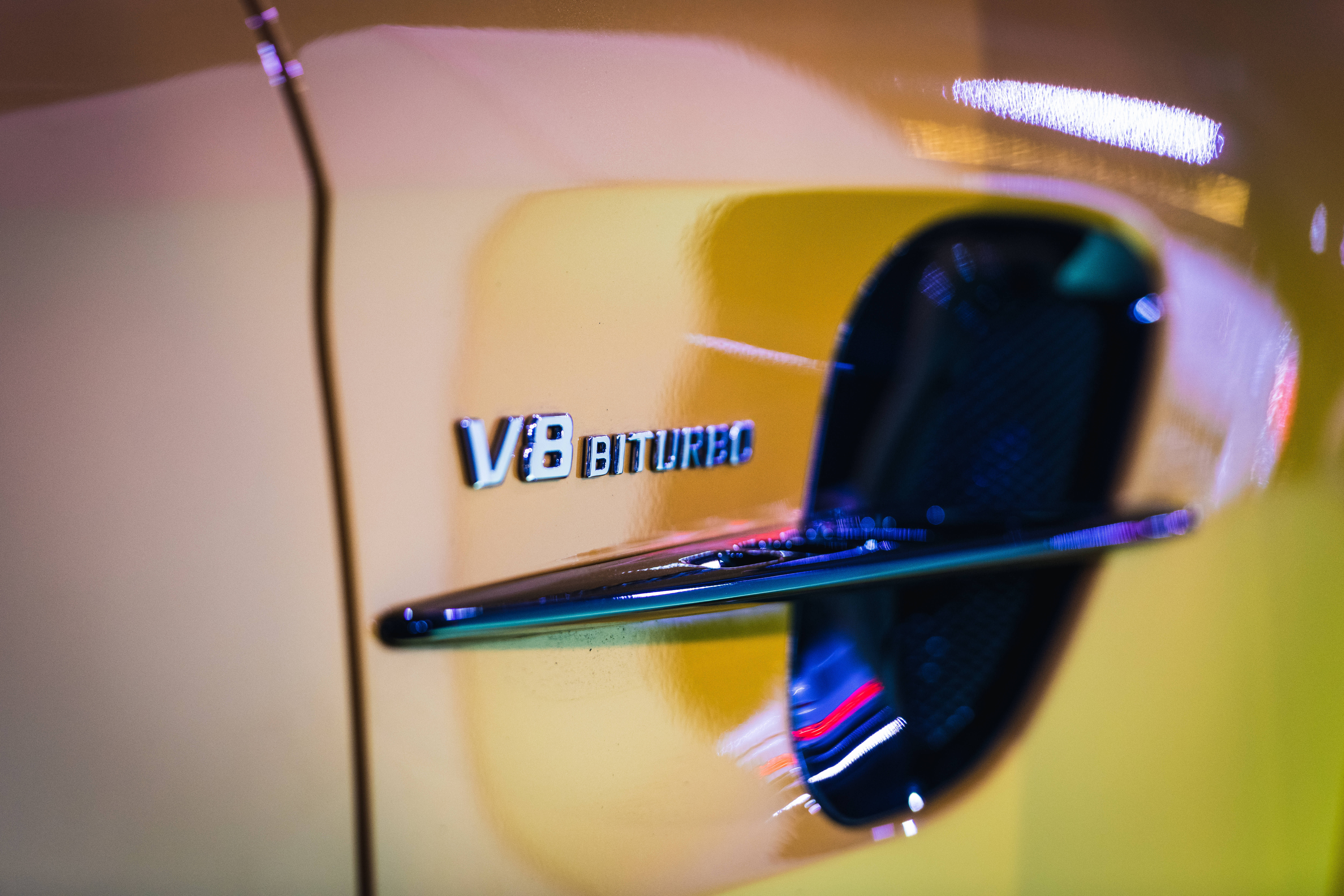 Mercedes Amg 桌布 壁紙 — 黃色車的V8 BITURBO徽章特寫，背景有模糊反射和霓虹燈。