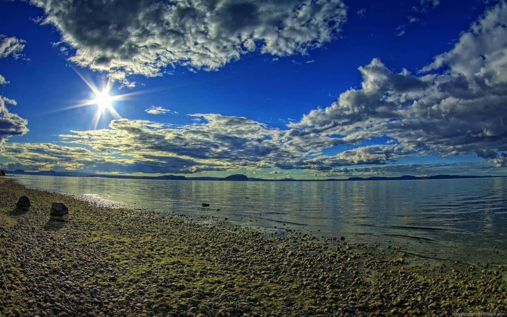Sunlight 桌布 壁紙 — 黃金時分，陽光穿透戲劇性的雲層，照耀在平靜的海面與卵石灘交匯之處。