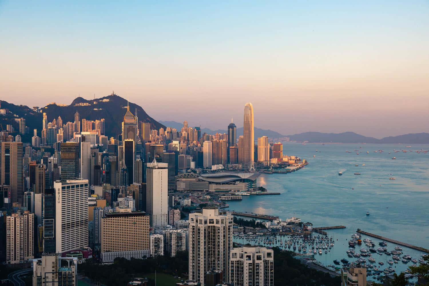 香港 桌布 壁紙 — 黃金時刻以溫暖的光芒沐浴香港的天際線，摩天大樓在滿是船隻的港口旁聳立。