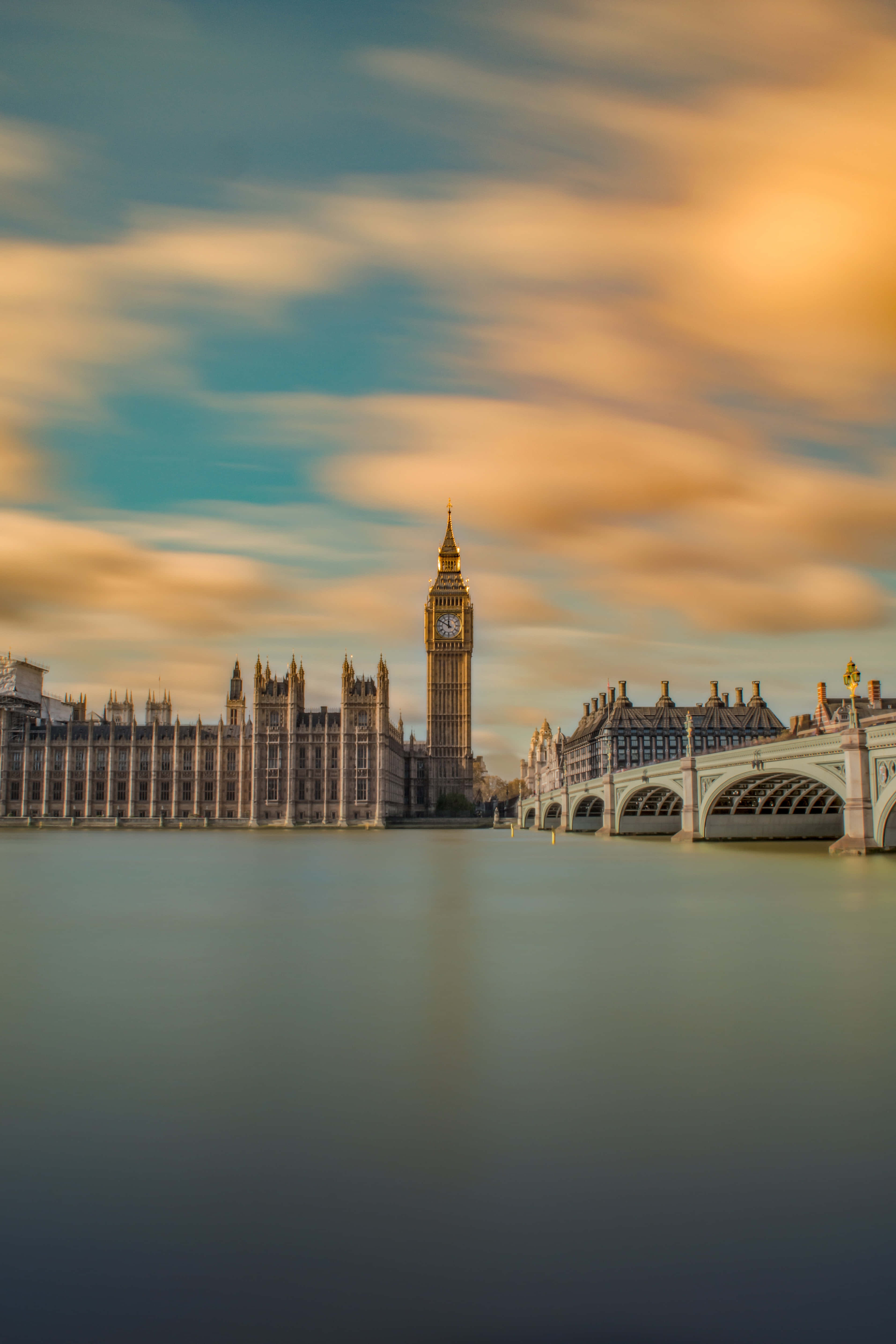 London iPhone 桌布 壁紙 — 黃金時刻的光芒以大笨鐘和西敏橋沐浴在溫暖色調中，雲彩在平靜的泰晤士河水面上劃過天空。
