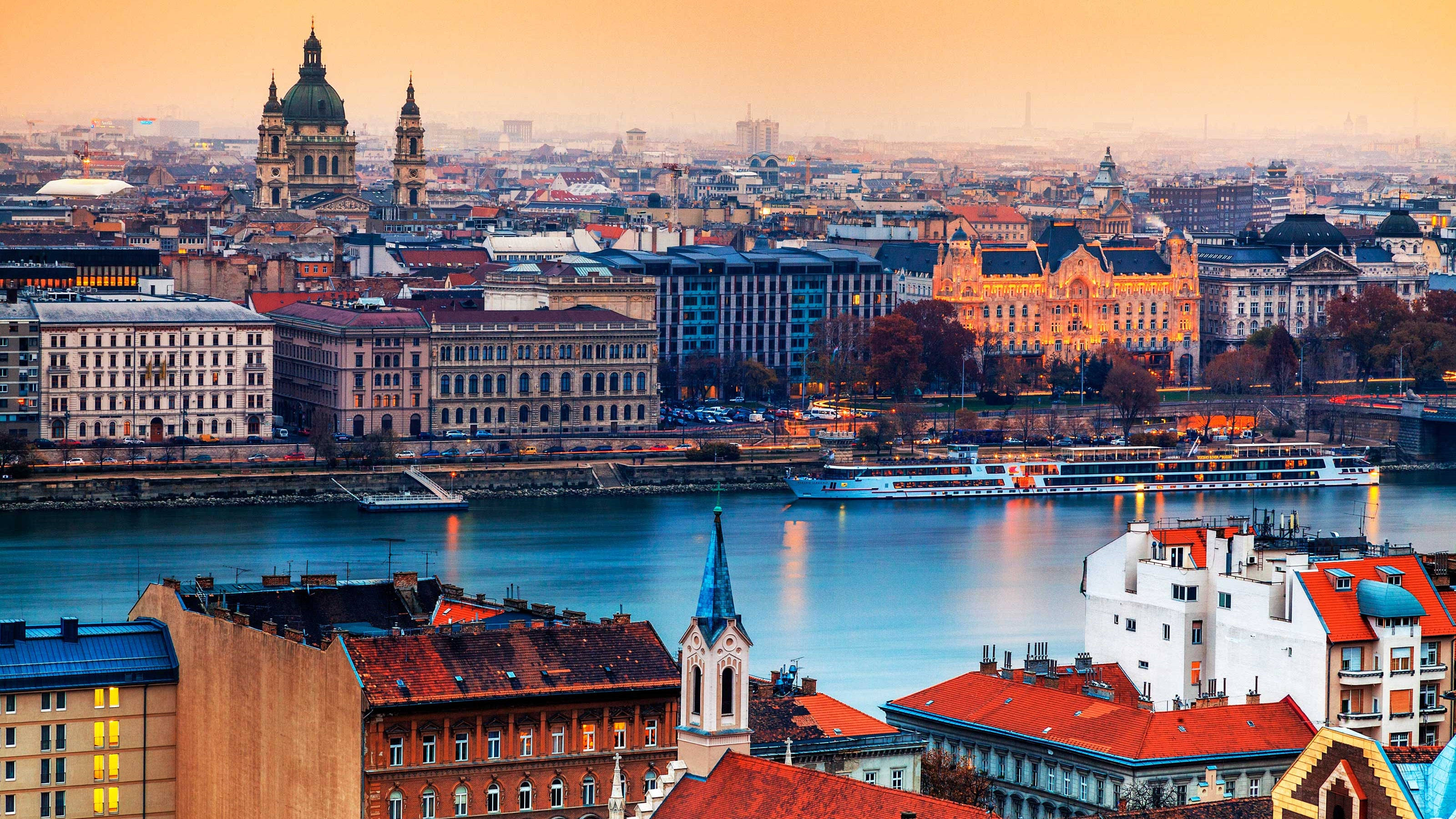 Hungary 桌布 壁紙 — 黃金時刻的光芒以溫暖的光線沐浴著布達佩斯的歷史天際線和多瑙河，河船在燈火通明的建築旁滑行。