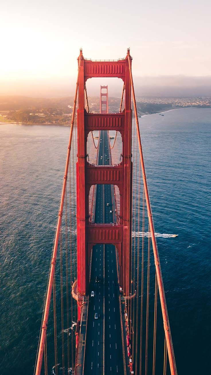 san francisco iPhone 桌布 壁紙 — 黃金時段，標誌性的紅色金門大橋橫跨深藍色水域的鳥瞰圖，遠方有山丘。