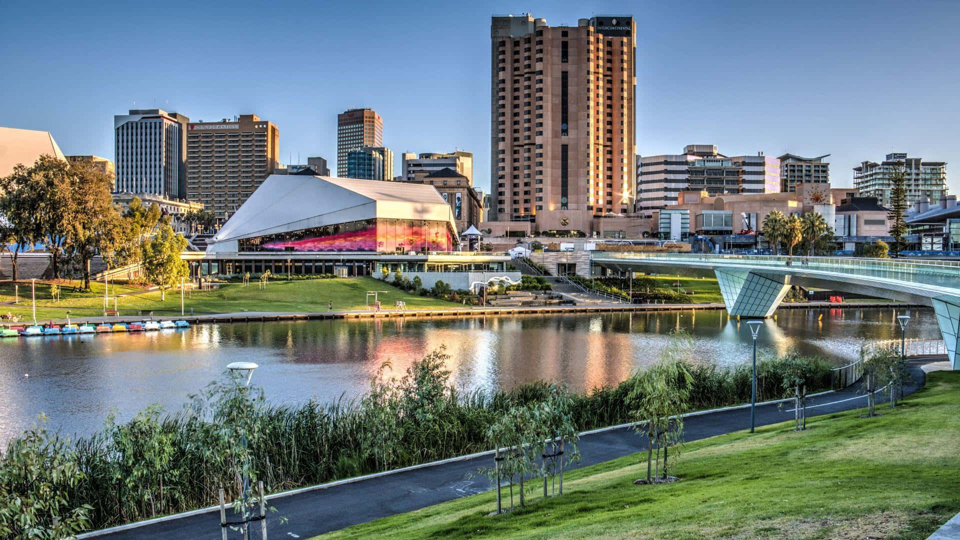 Adelaide 桌布 壁紙 — 黃金時段的現代城市景觀，玻璃橋橫跨平靜的河流，未來主義建築倒映在水中。