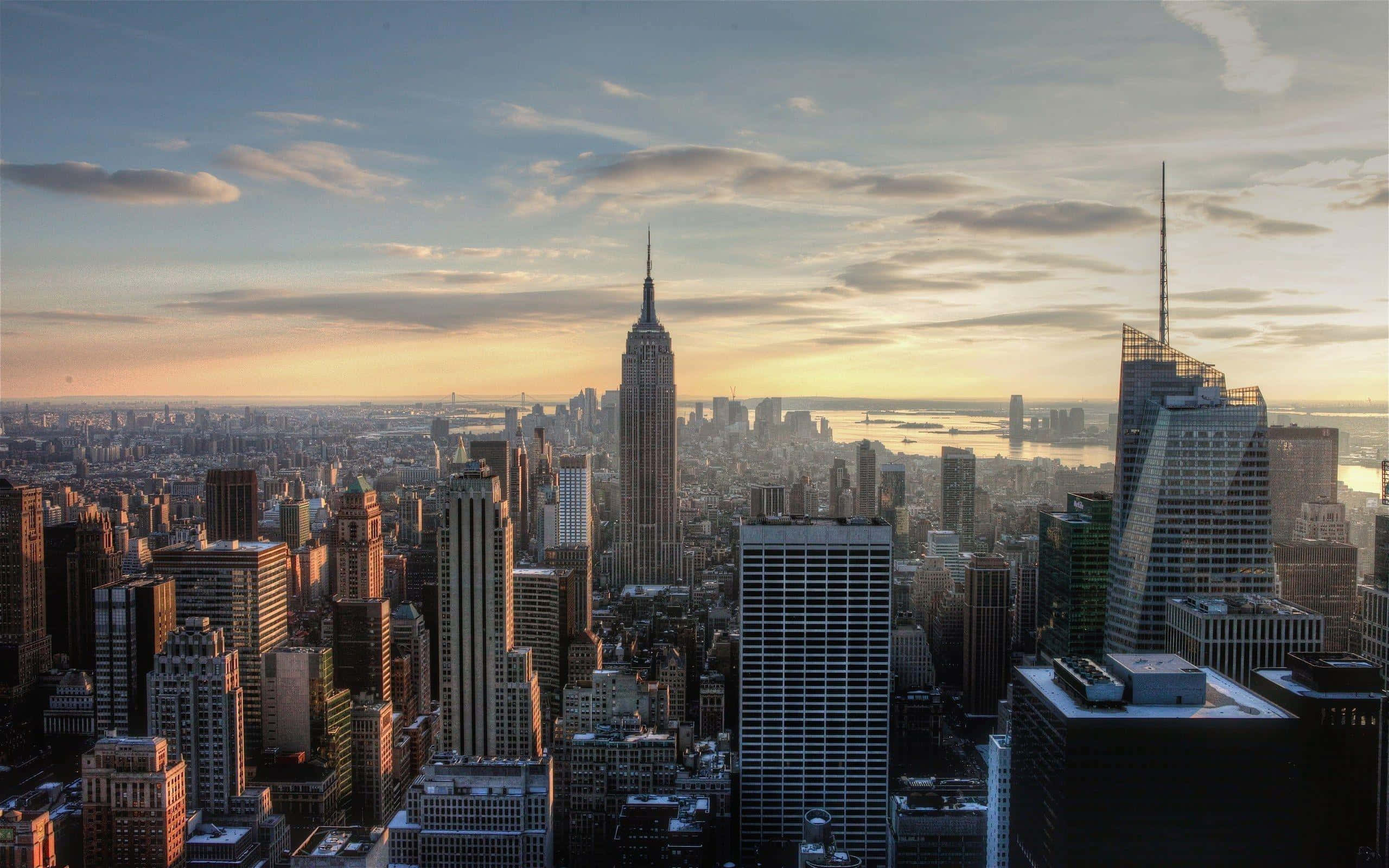 New York 桌布 壁紙 — 黃金時段的陽光以溫暖色調沐浴著標誌性的紐約市天際線，雲朵在高聳的摩天大樓上方飄移。