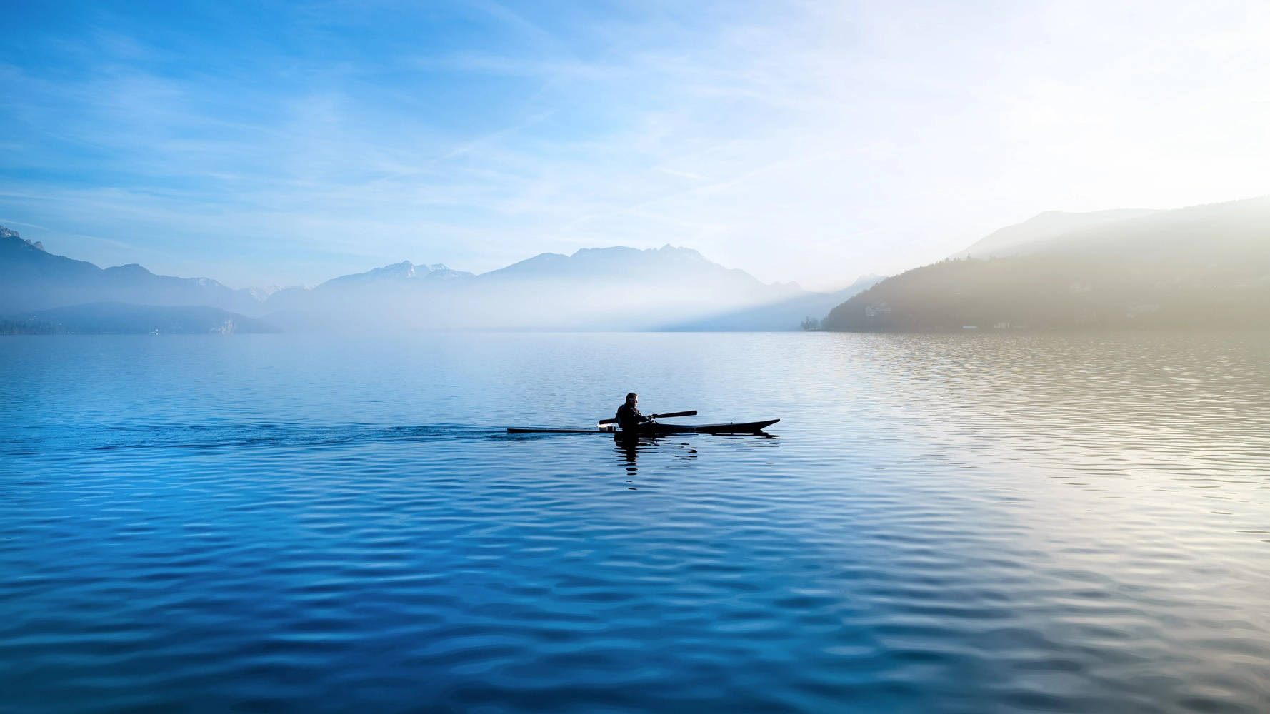 Rowing 桌布 壁紙 — 黎明時分，一名孤獨的划船者在柔和的藍天下滑過霧氣瀰漫的山間湖泊。